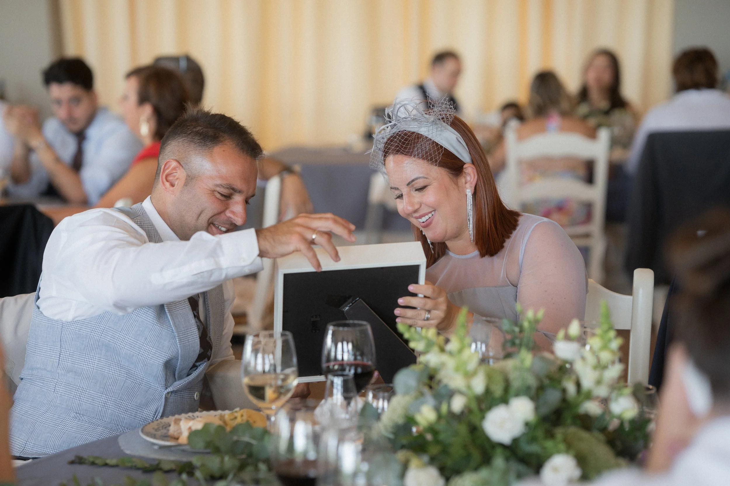 Una pareja en una boda recibe un regalo de bodas, mirando un álbum de fotos con una sonrisa, rodeados de otros invitados y decoraciones florales.