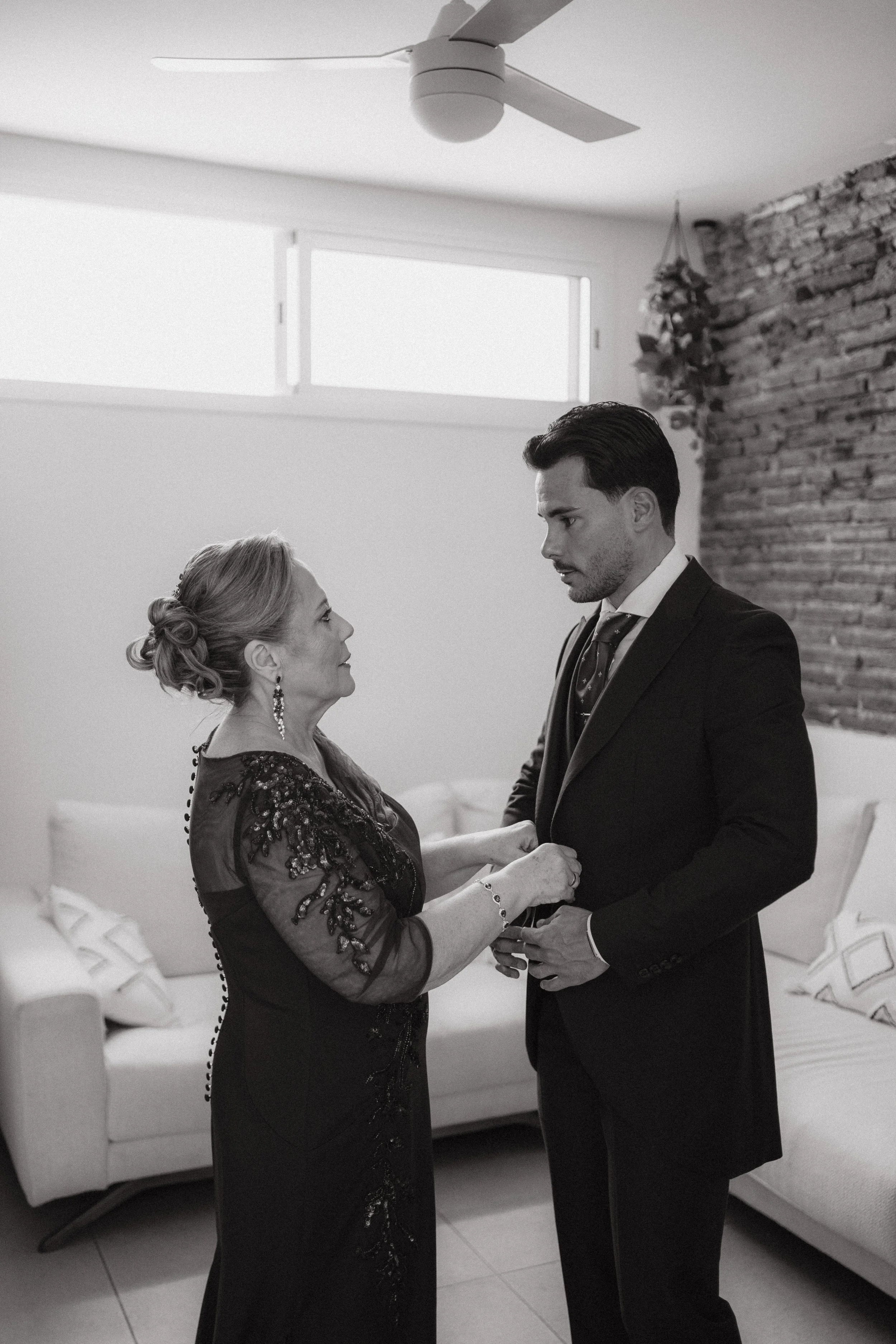 Una mujer mayor y un hombre joven en un salón, la mujer le ajusta la chaqueta al hombre mientras los miran. La escena es en blanco y negro.