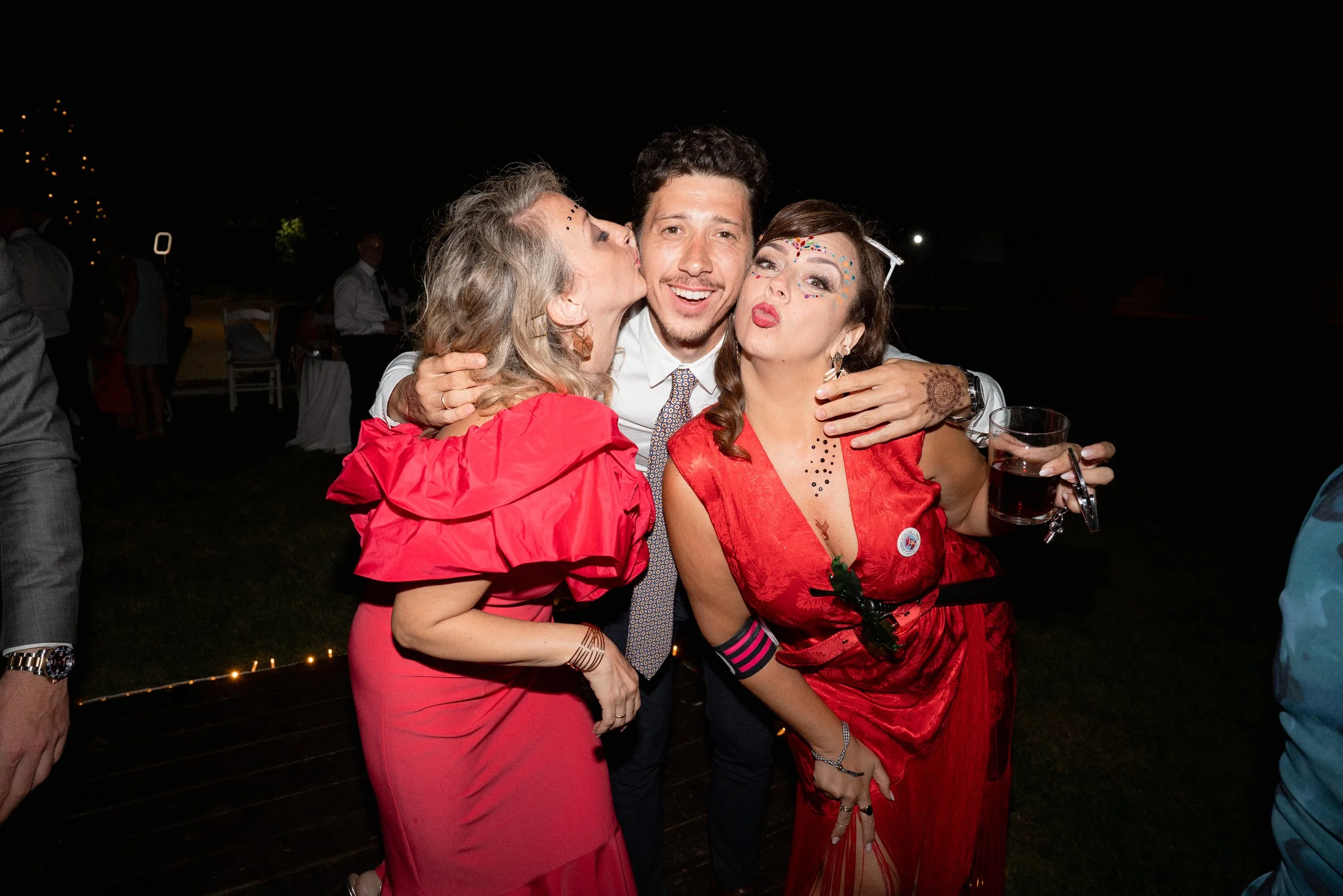 Tres personas en una fiesta nocturna, dos mujeres con maquillaje y ropa rojos, un hombre en medio sonriendo, abrazándose y posando para la cámara.