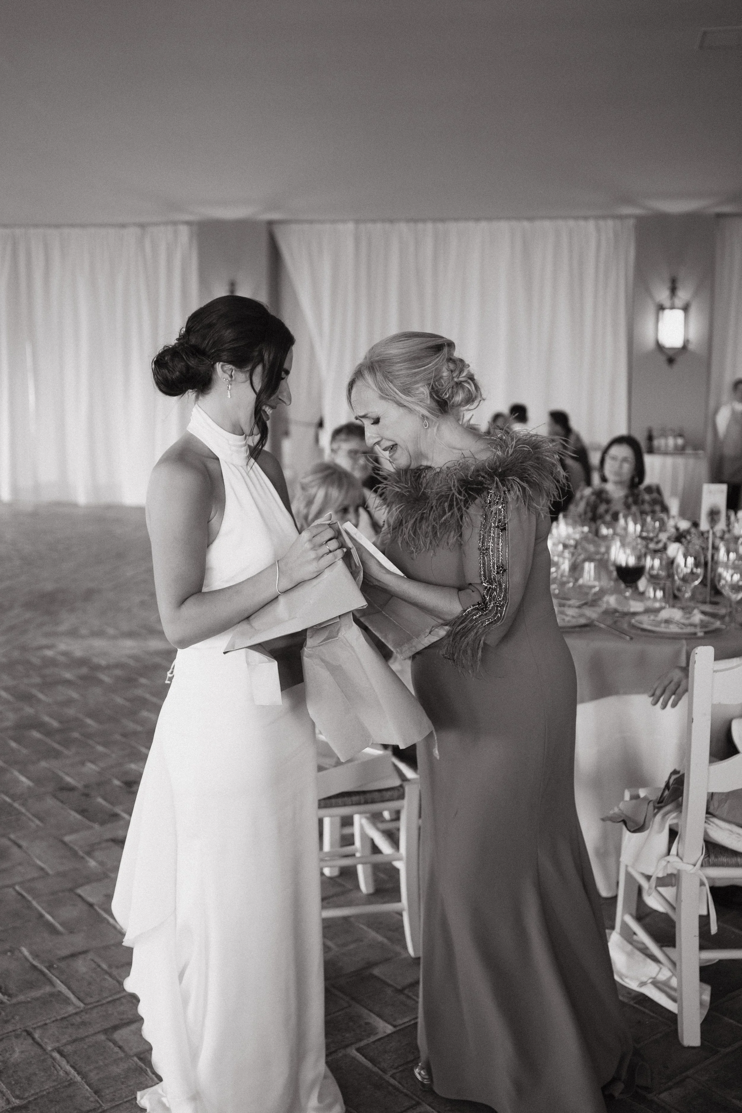 Dos mujeres en un evento, una vestida con vestido largo blanco y la otra con vestido largo con detalles de plumas en los hombros, parecen compartir un momento emotivo y alegre, con otras personas en el fondo en un espacio decorado para una celebració