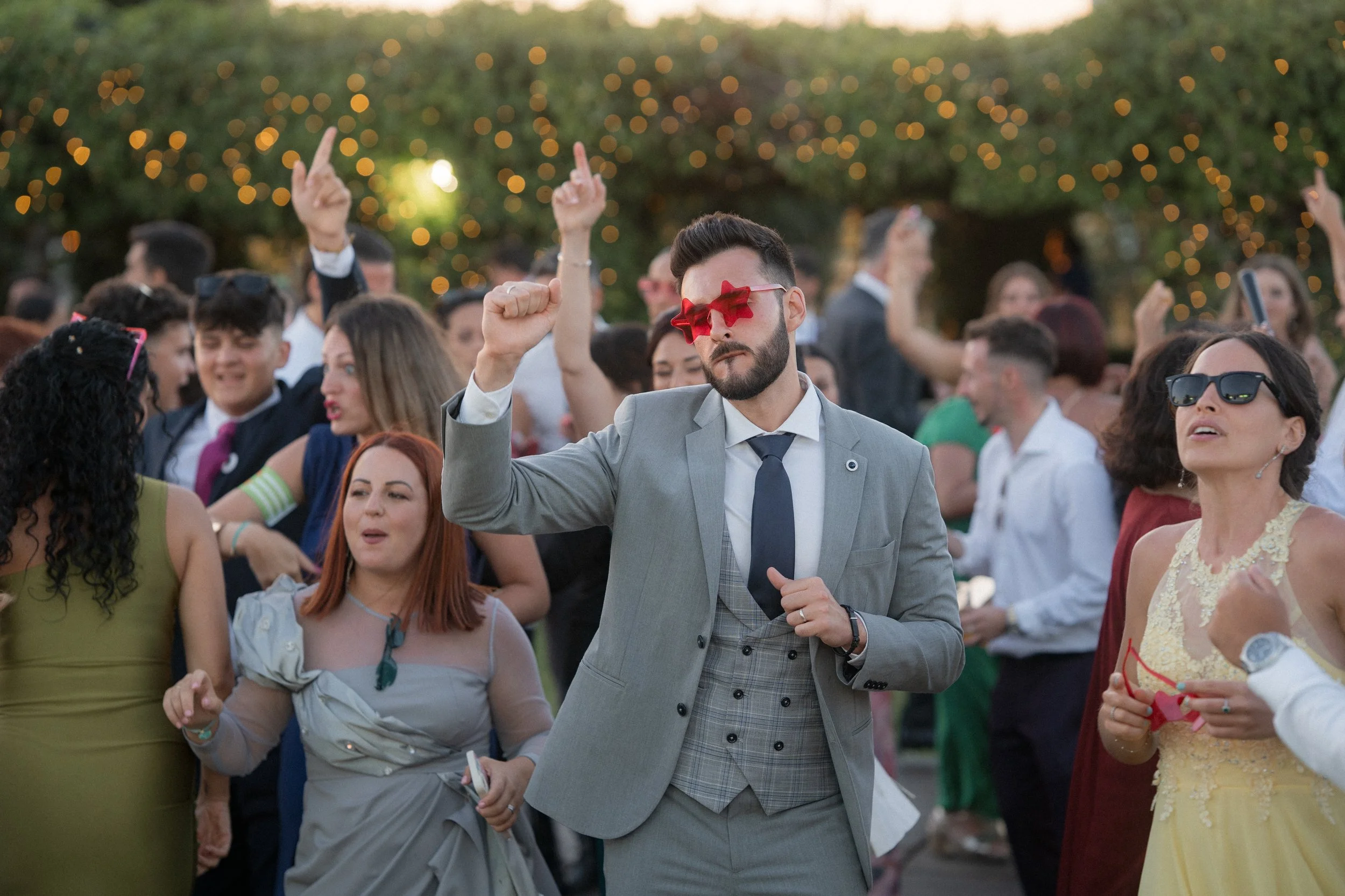 Personas bailando en una fiesta al aire libre con luces en el fondo, algunos usan gafas divertidas y ropa elegante.