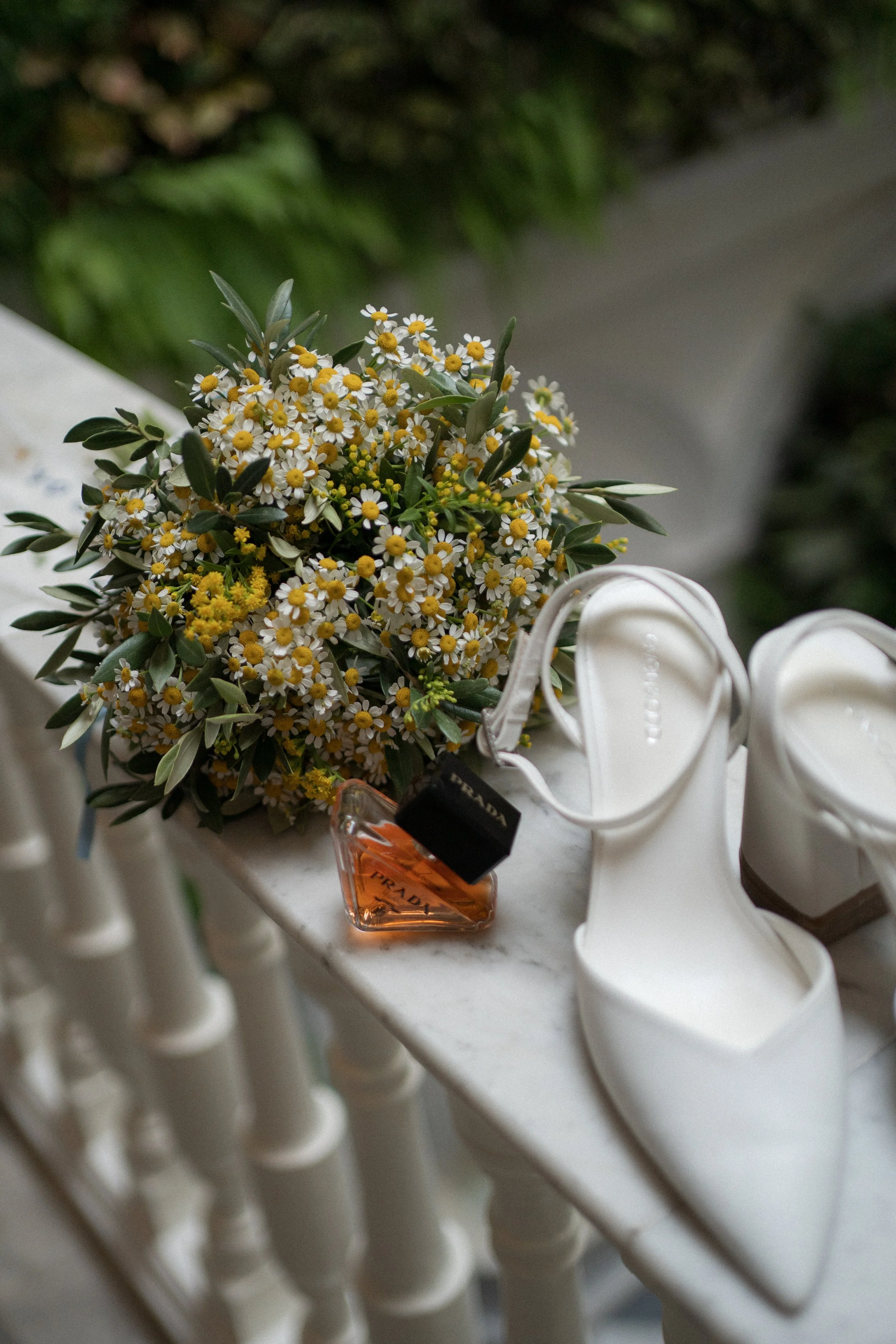 Zapatos blancos de tacón alto, ramo de flores pequeñas y blancas con amarillos, y perfume rosa en una mesa de mármol.