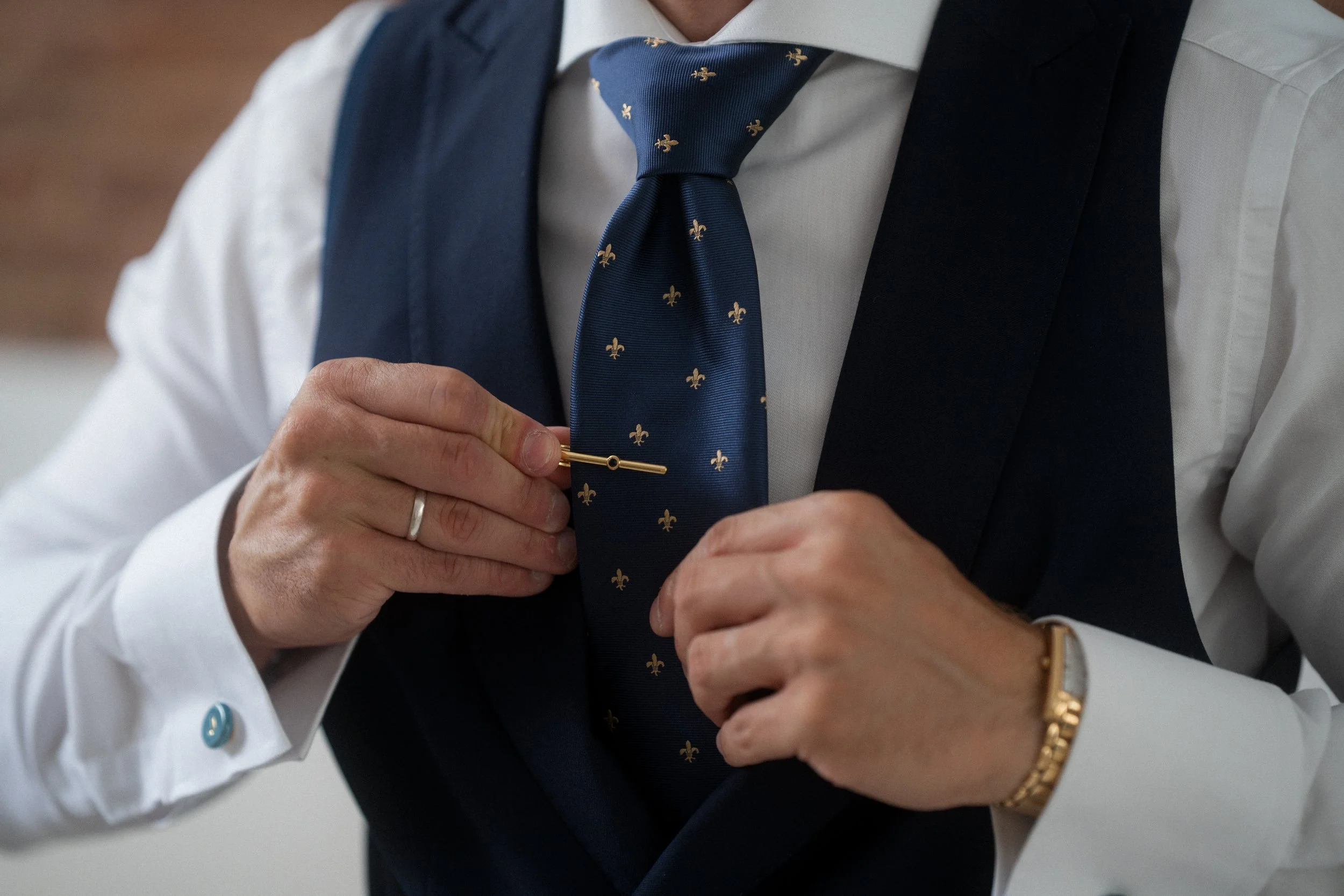 Persona arreglándose la corbata con un alfiler en un atuendo formal, llevando una camisa blanca, un saco oscuro y un reloj dorado.