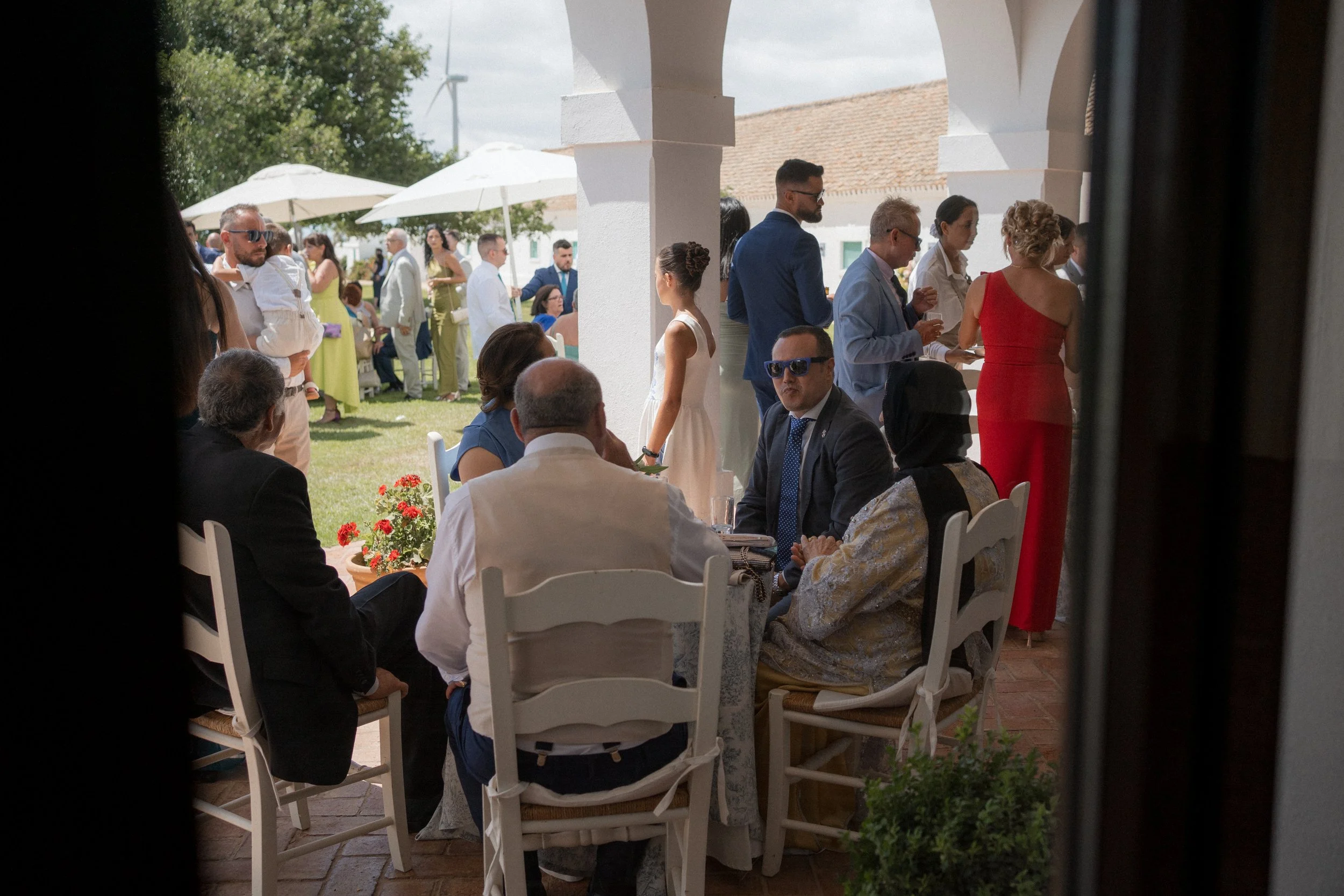 Personas reunidas en los aperitivos de una boda al aire libre en dehesa bolaños, algunas sentadas en una mesa y otras de pie, en un ambiente con sombrillas y árboles.