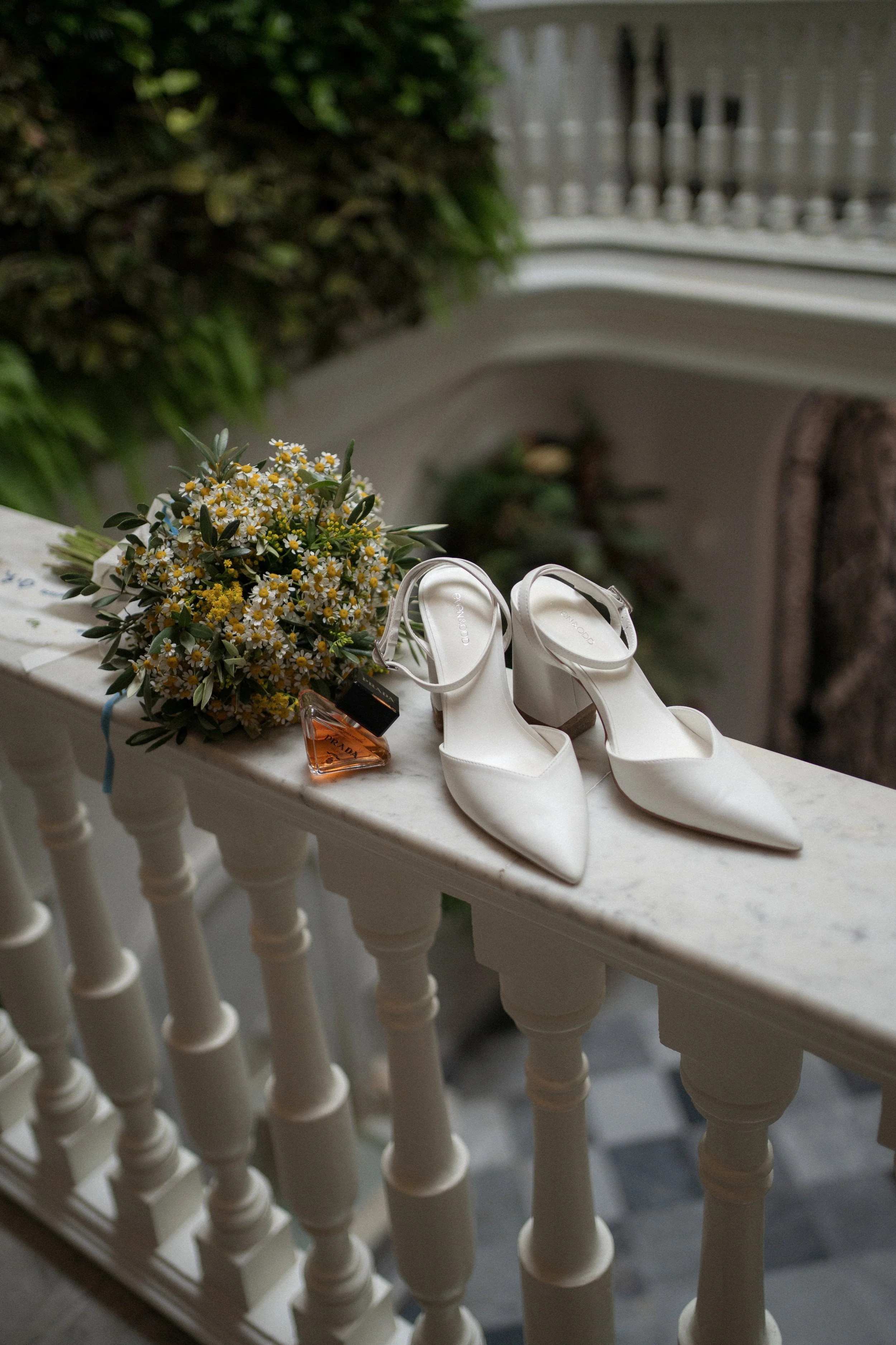Zapatos blancos de tacón con lazo, ramo de flores y perfume en una barandilla de mármol en un interior decorado con plantas.