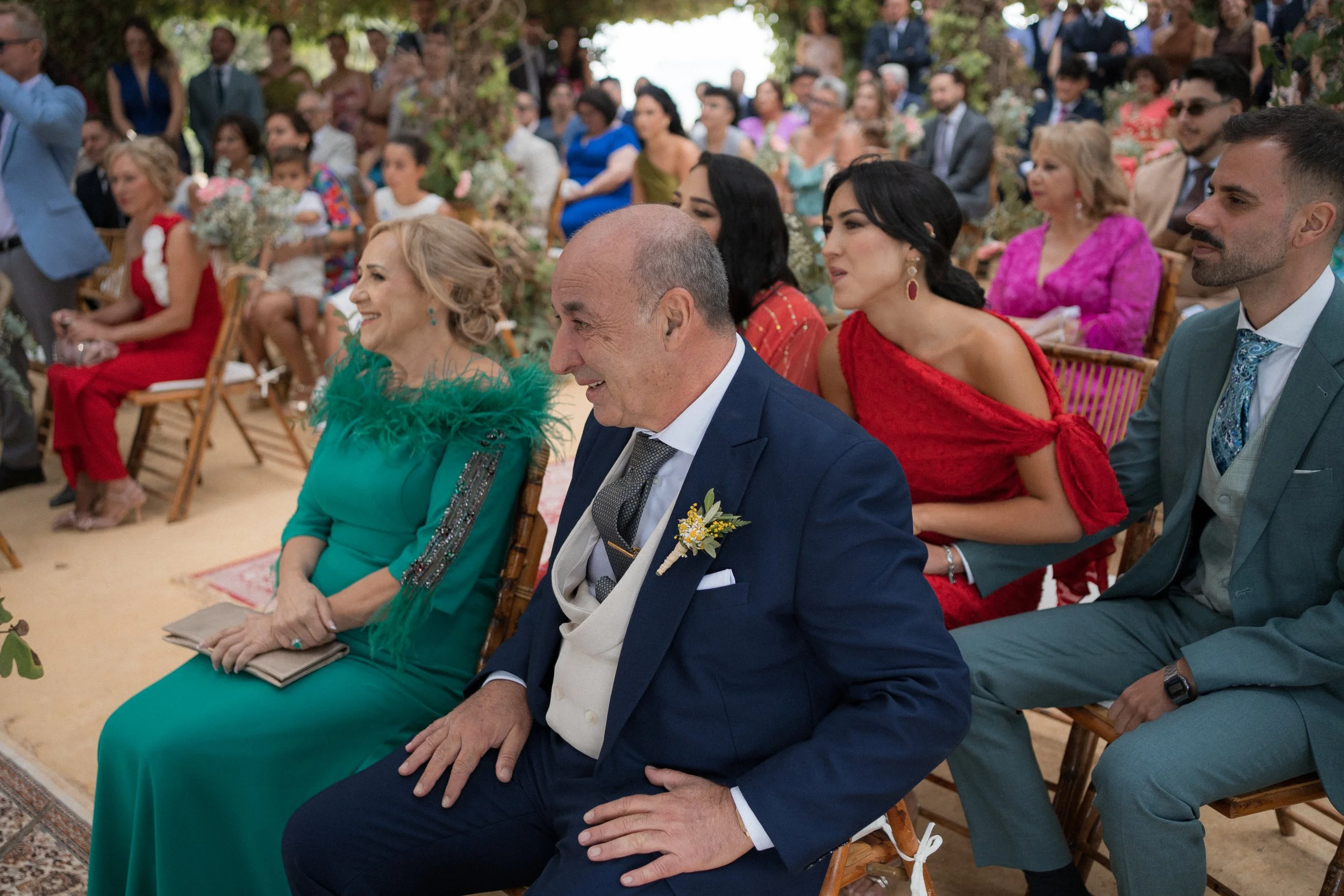 Grupo de personas sentadas en una ceremonia, algunas con trajes elegantes y coloridos, rodeadas de decoraciones florales.