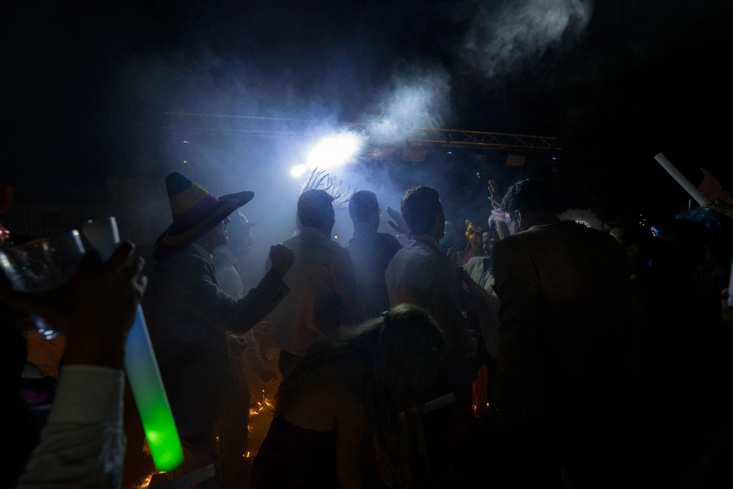 Gente disfrutando en una fiesta nocturna con luces y humo de fondo, algunas personas con sombreros y accesorios que pertenecen a la hora loca de una boda.
