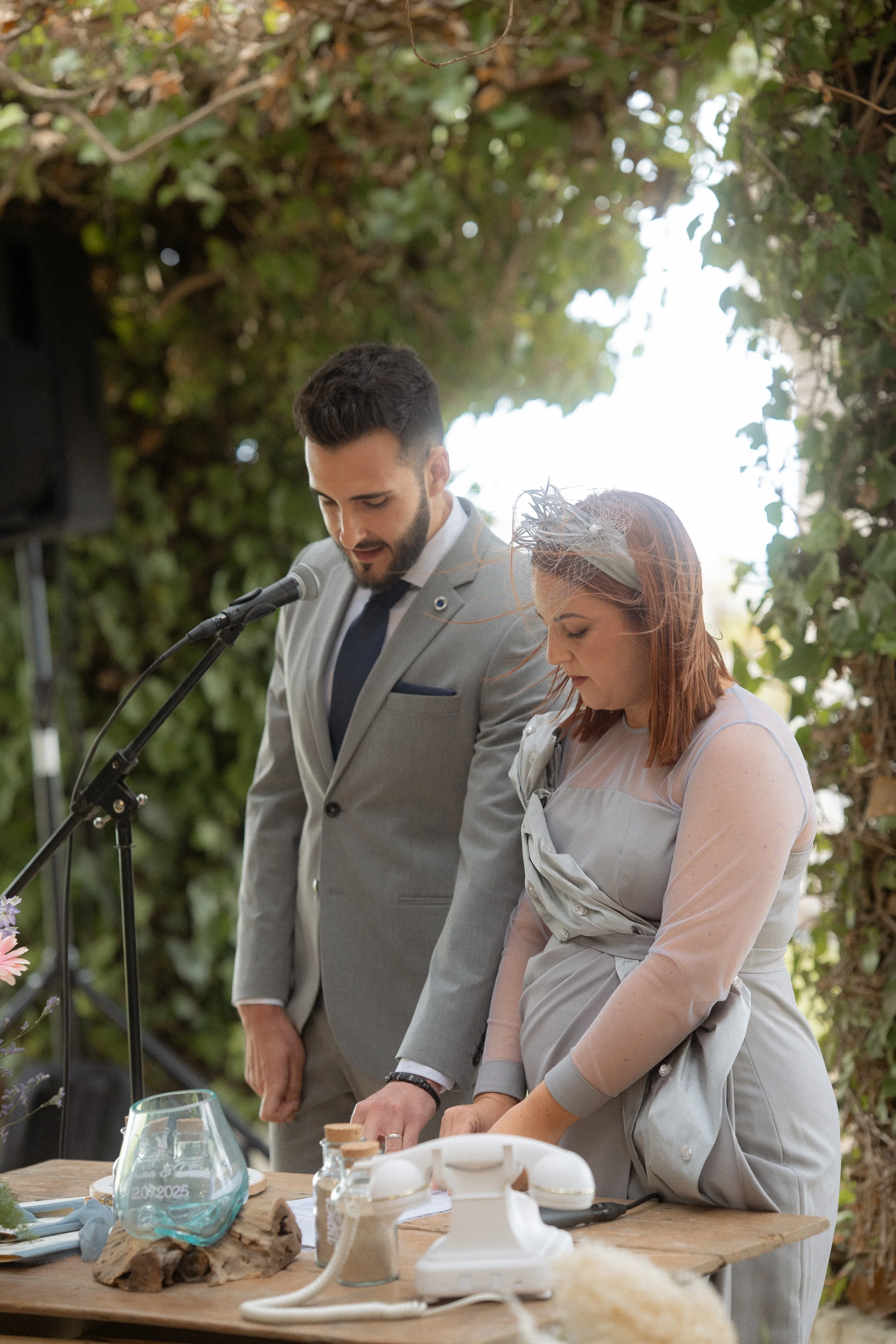 Oficiantes de boda, leen en la ceremonia, con un fondo de vegetación verde y un escritorio con elementos decorativos y un teléfono antiguo.
