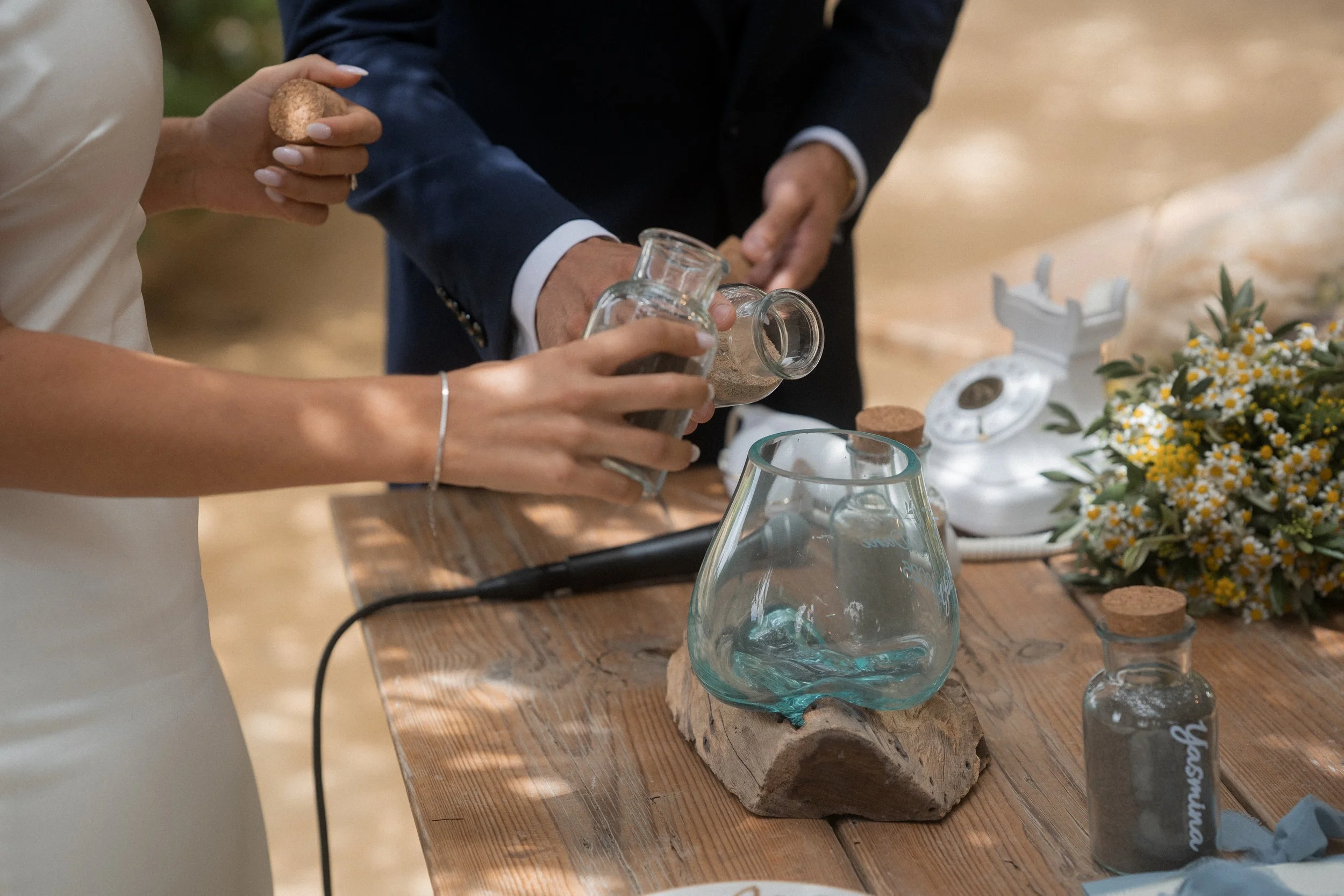 Dos personas en una ceremonia, una de ellas sosteniendo un frasco pequeño y la otra ayudando a verter polvo en un vaso de cristal puesto sobre una base de madera, junto a una floristería de flores pequeñas blancas y amarillas y un teléfono antiguo de