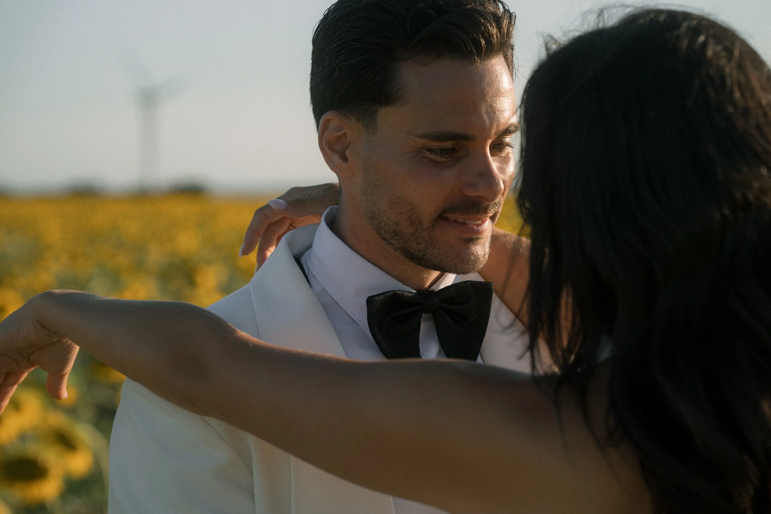 Una pareja bailando en un campo de girasoles, él viste un esmoquin con pajarita, ambos parecen felices y románticos.