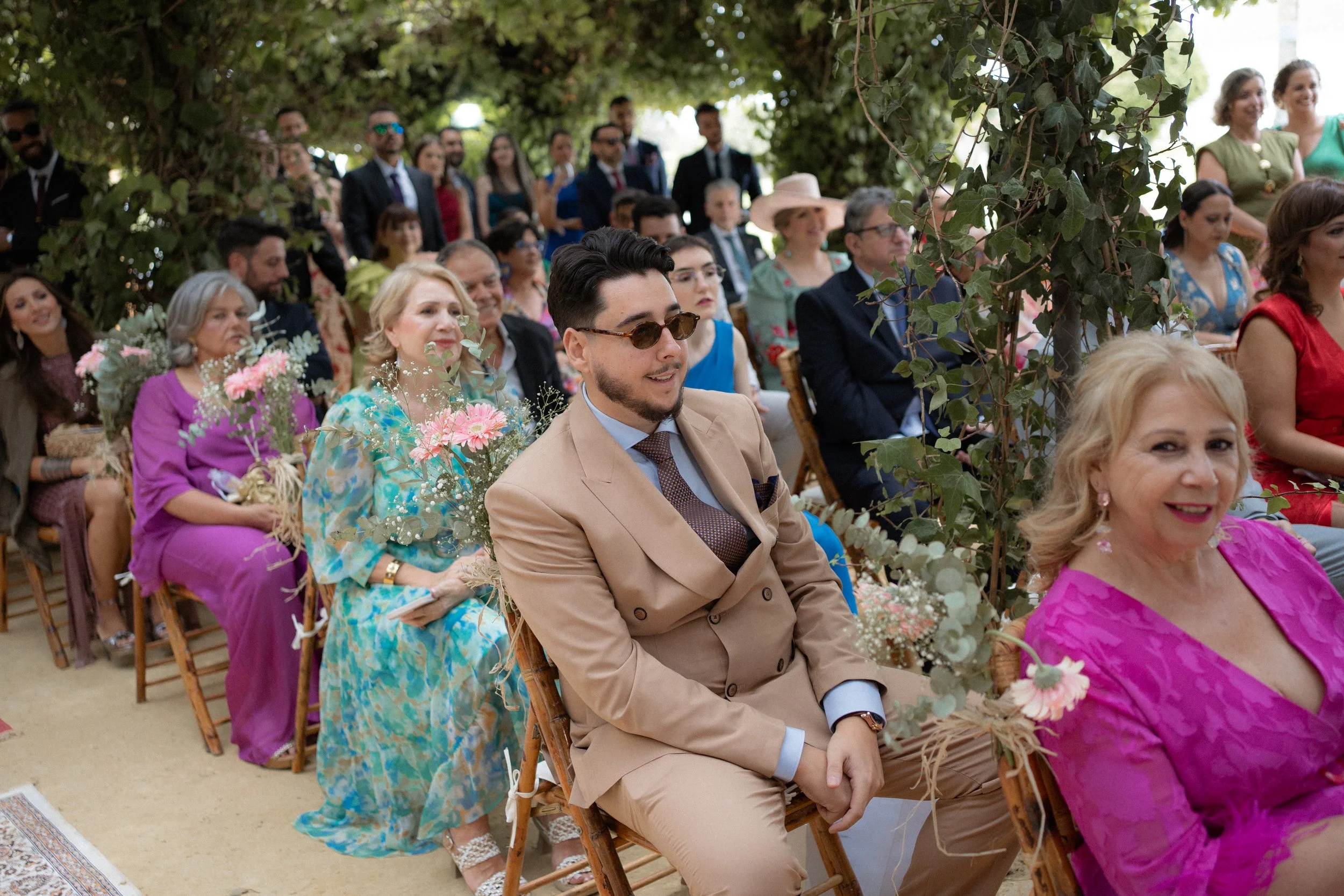Hombres y mujeres sentados en sillas, asistiendo a una ceremonia al aire libre, decorada con plantas y flores.