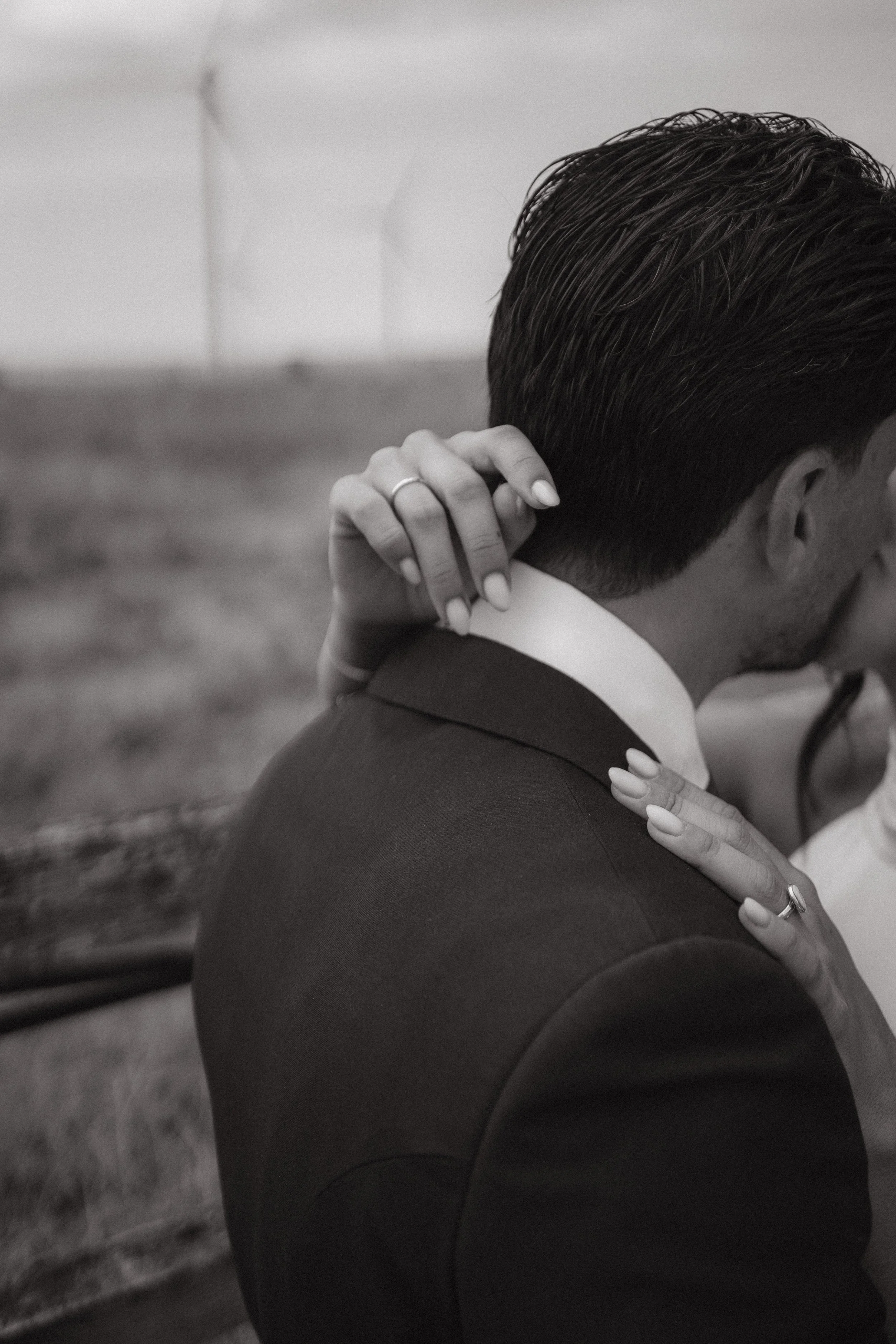 Un hombre y una mujer en un abrazo, la mujer tiene la mano en el cuello del hombre y lleva un anillo en el dedo, fondo con molinos de viento en un campo.