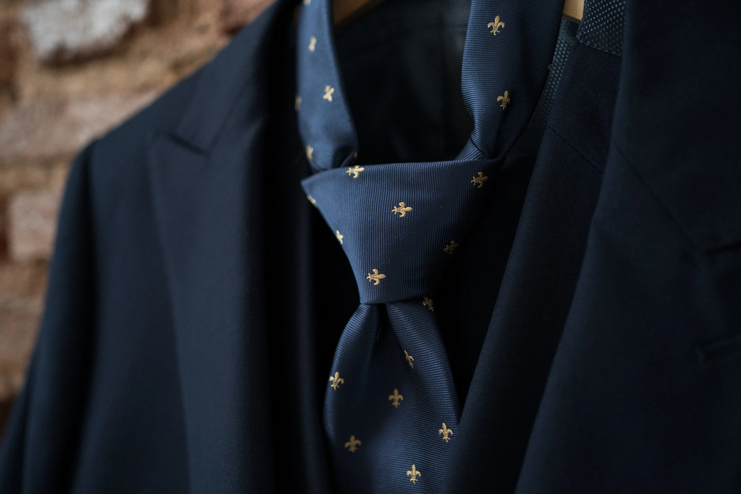 Corbata azul marino con pequeños olivos dorados, puesta con traje oscuro y fondo de ladrillos.