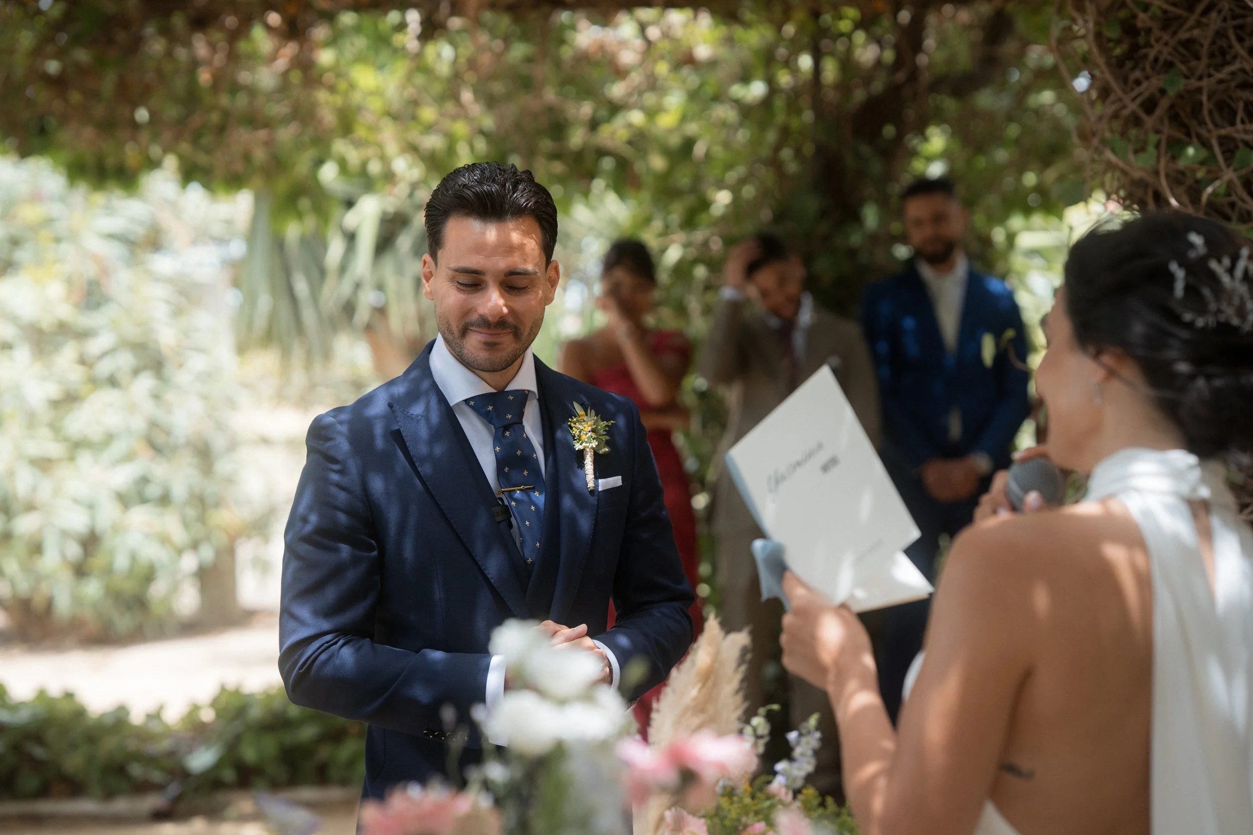 Un hombre y una mujer en una ceremonia de boda al aire libre, con el hombre vistiendo un traje azul y la mujer con un vestido blanco y tiara, rodeados de árboles y flores.