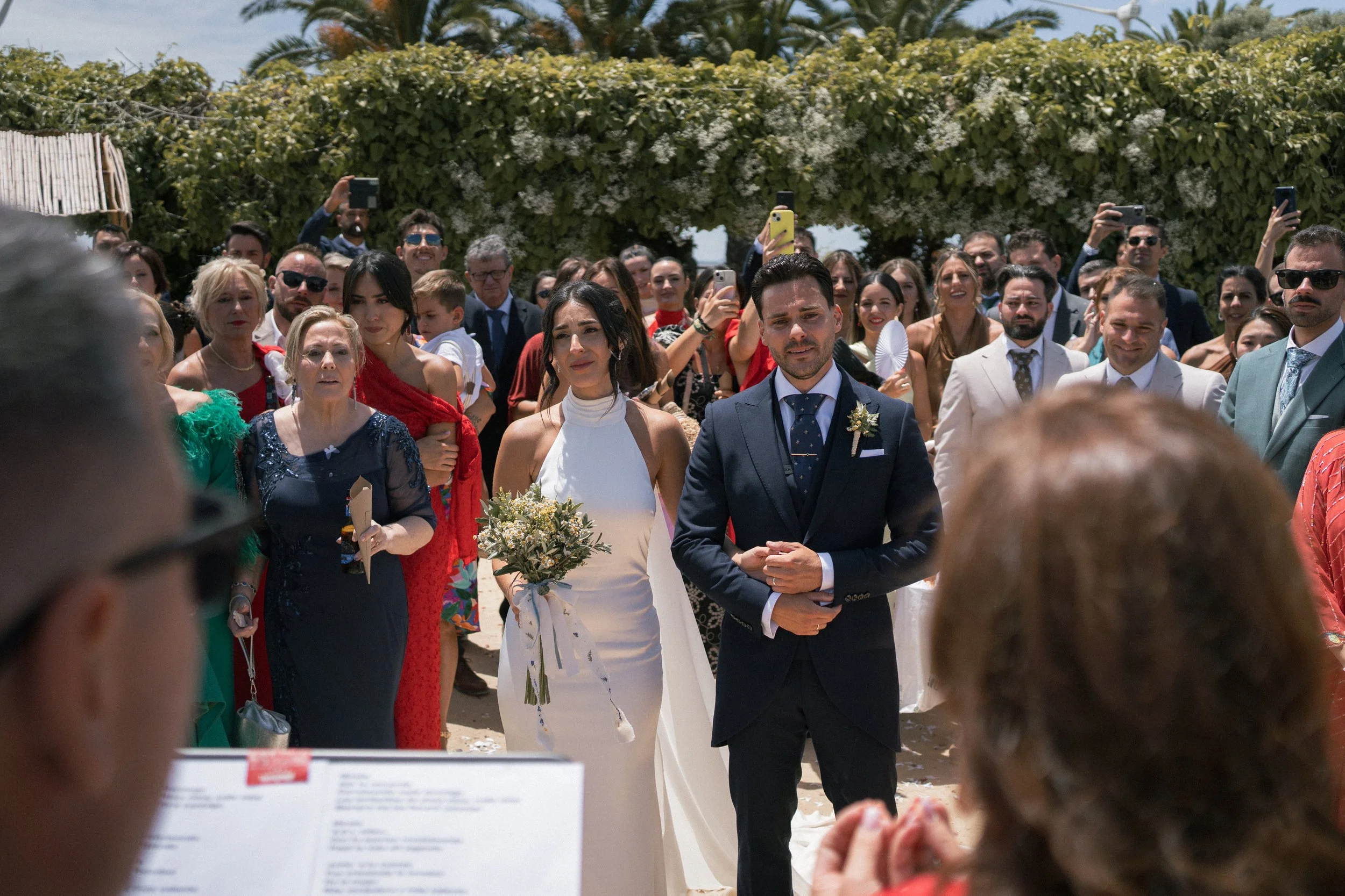 Pareja de novios en su boda, rodeados de invitados, al aire libre con cielo despejado y vegetación de palmeras y plantas.