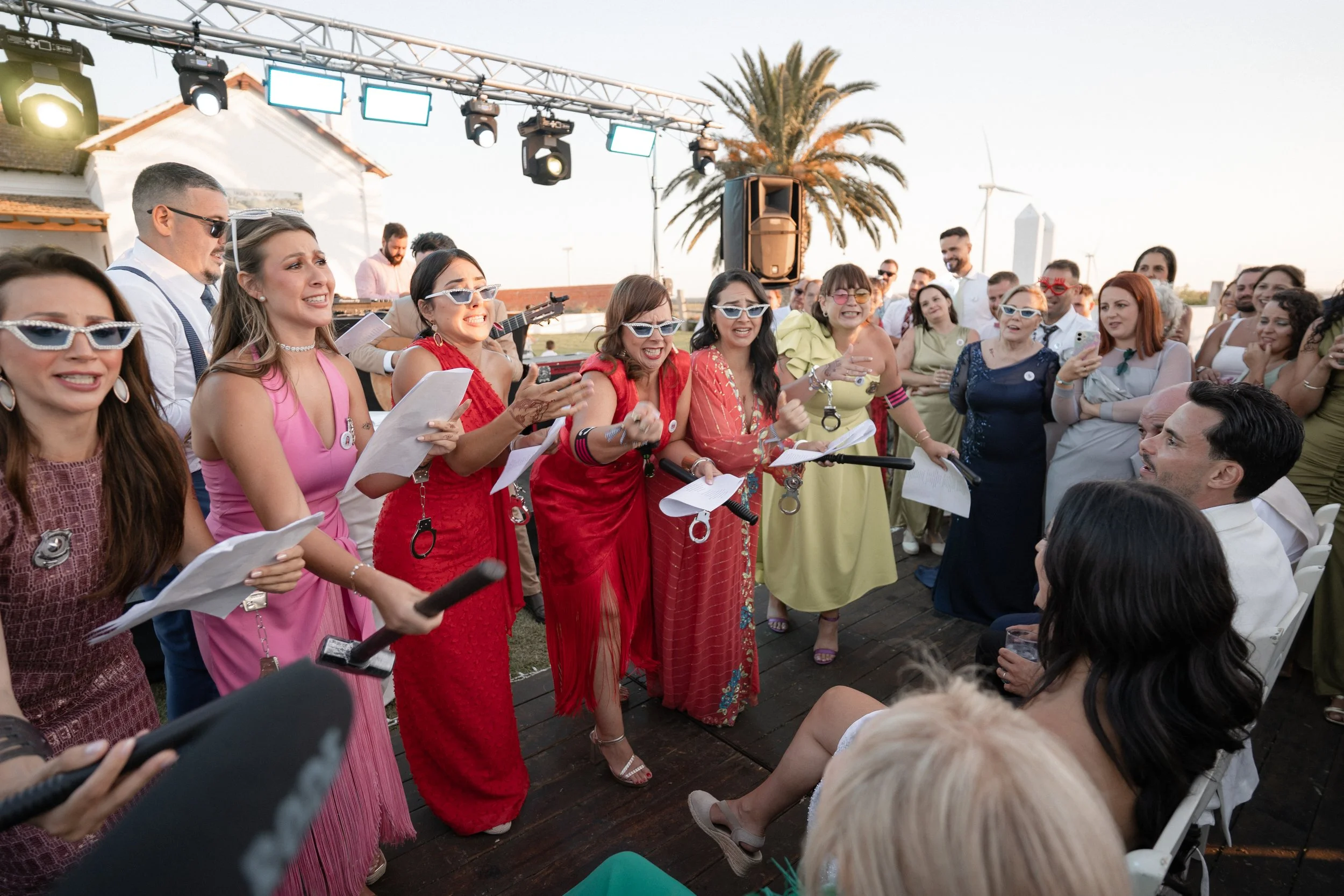 Un grupo de personas en una celebración al aire libre, con algunas cantando y otras disfrutando de la compañía, en un ambiente festivo y soleado con cadenas de luces y un fondo con palmeras y un viento eólico.