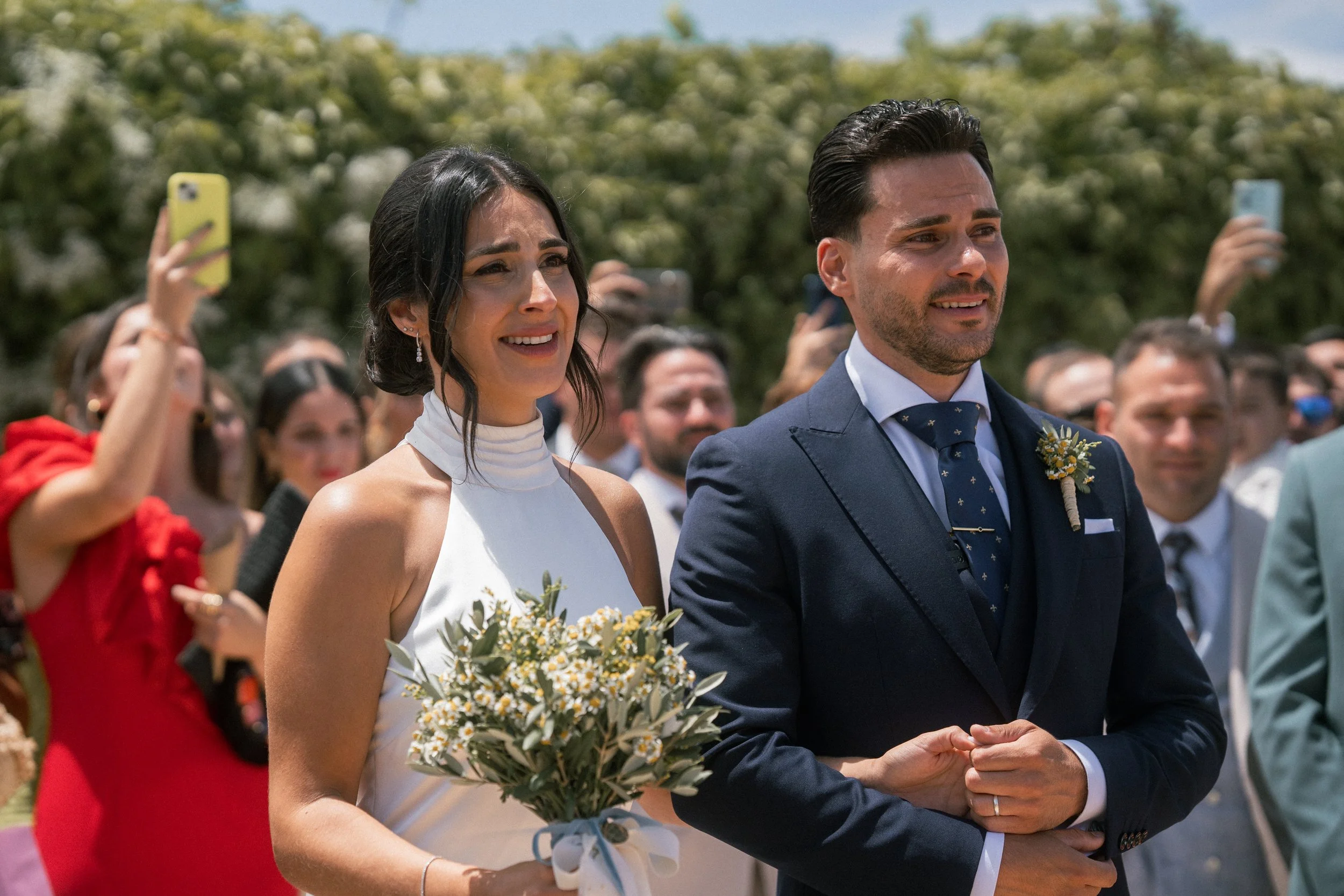 Pareja de boda, la novia con un ramo de flores y el novio con traje oscuro, en una ceremonia al aire libre, rodeados de invitados.