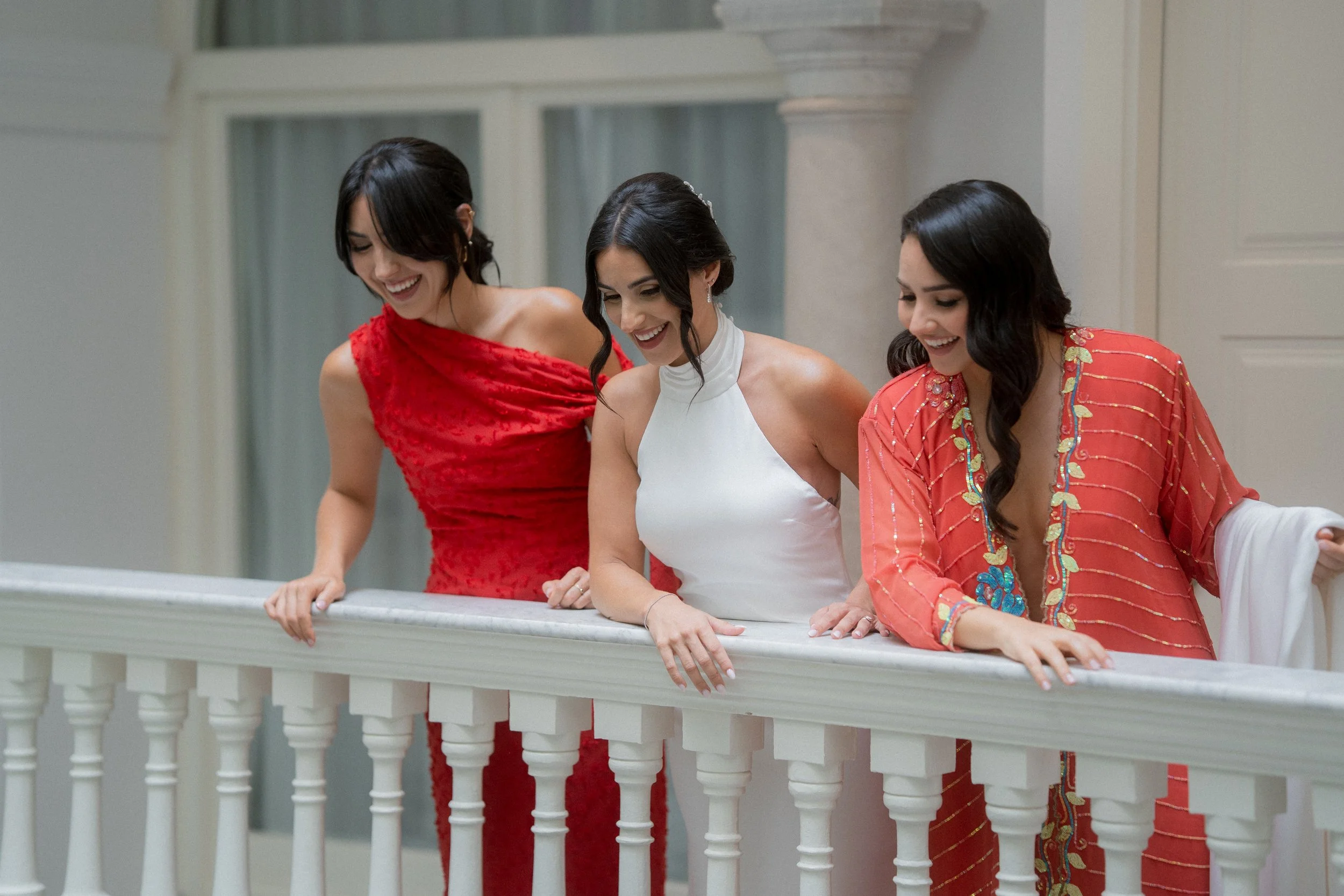 El mejor fotografo de bodas retrata a tres mujeres sonriendo y mirando hacia abajo desde una barandilla en un lugar elegante y bien decorado.