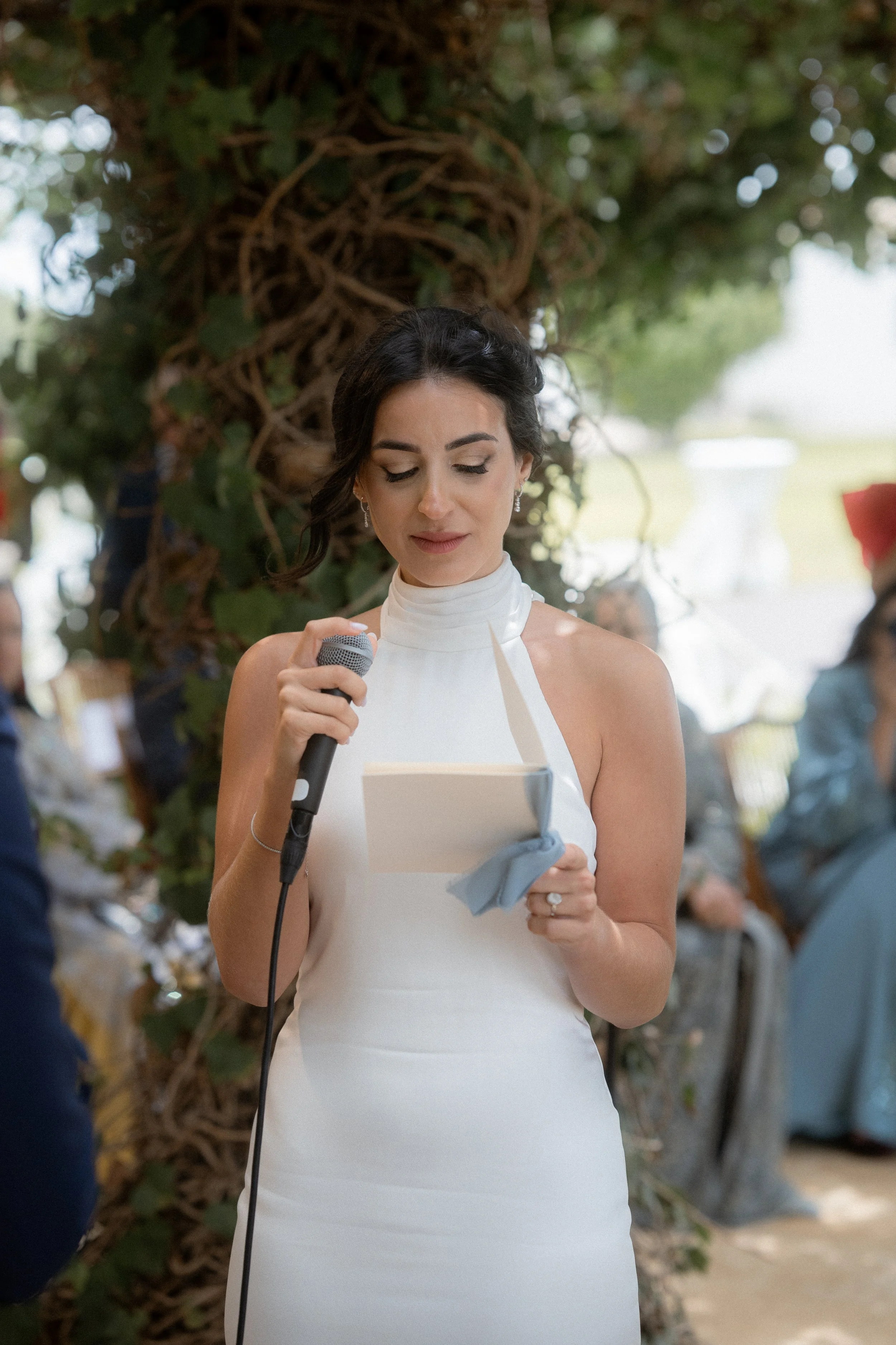 Mujer en vestido blanco leyendo un discurso con micrófono en un evento al aire libre, rodeada de naturaleza y en una ceremonia