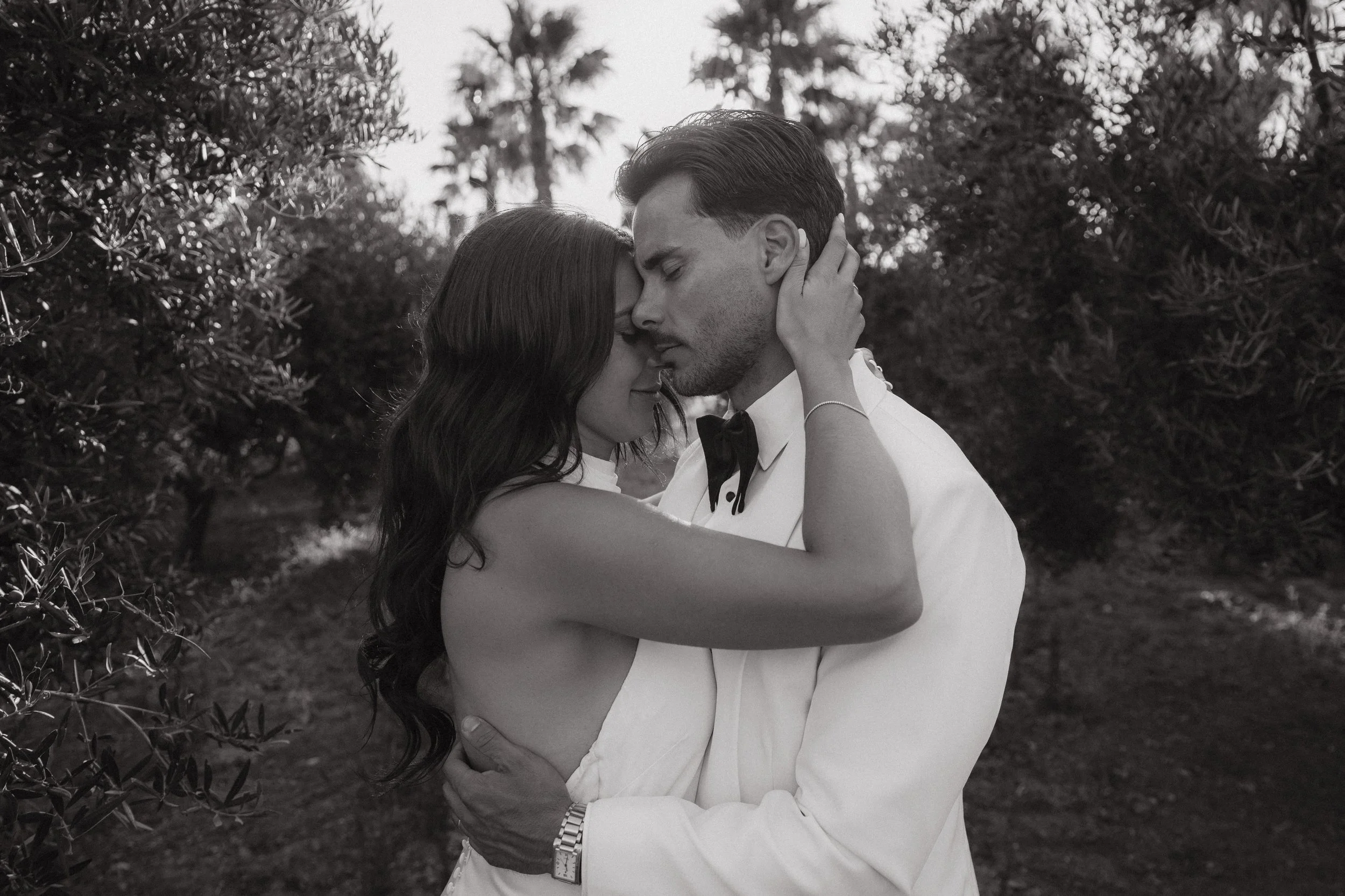 Una pareja vestida formalmente abrazándose y tocándose la frente en un entorno natural. La mujer viste un vestido sin tirantes y el hombre lleva un esmoquin con pajarita, en una escena en blanco y negro.