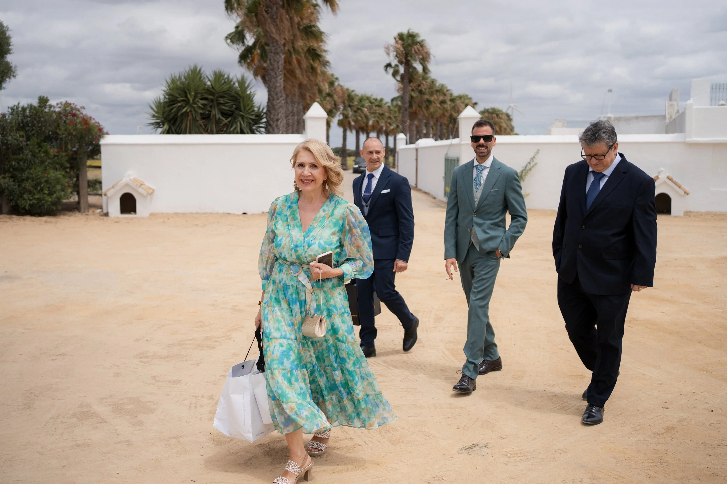 Grupo de cinco personas caminando en un camino de arena en un lugar con palmeras y paredes blancas, vestidos formalmente, en un día nublado.