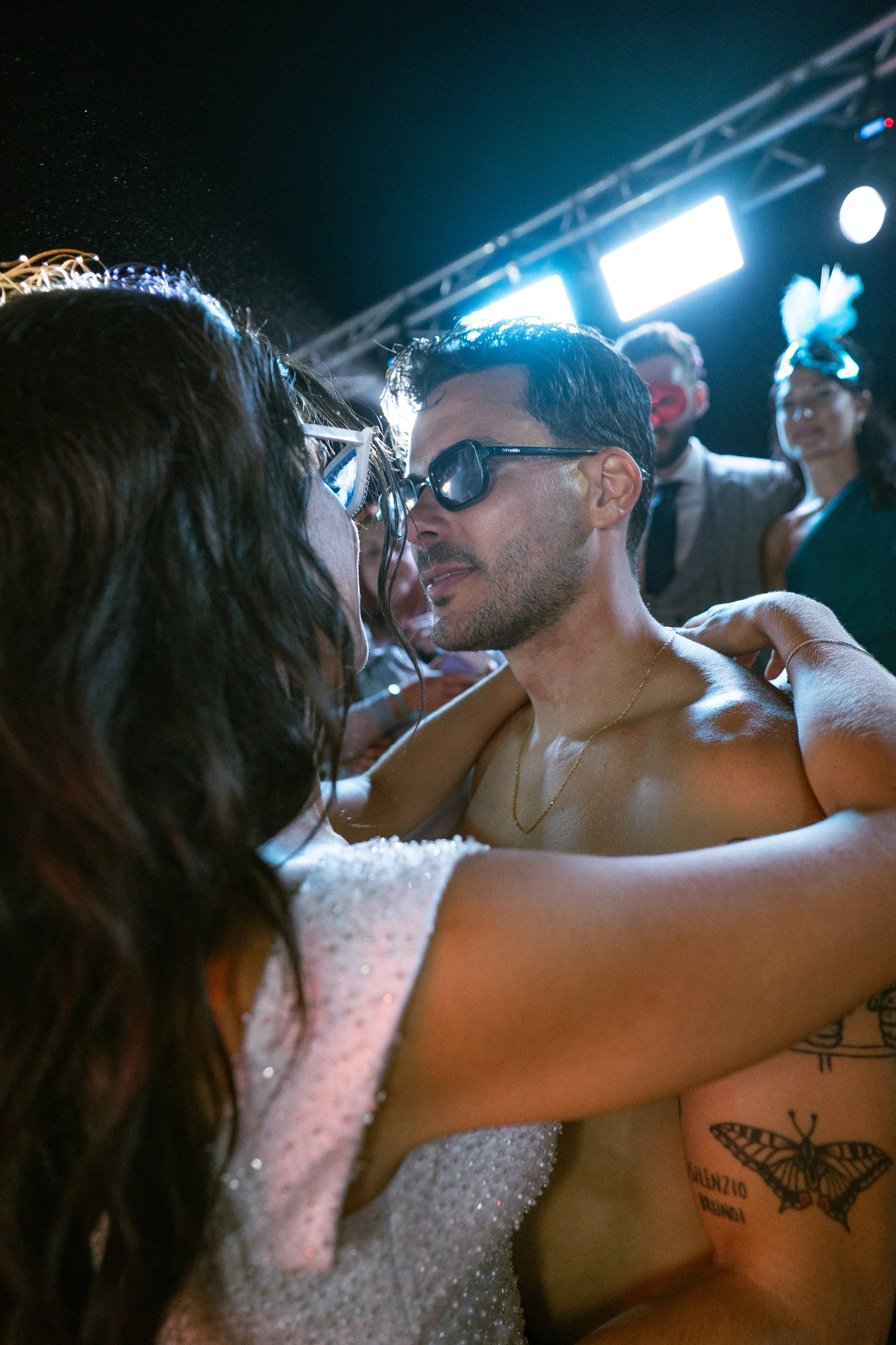 Pareja bailando en una fiesta con fondo de luces brillantes, rodeados de otros asistentes. La mujer tiene el cabello largo y usa un vestido brillante; el hombre lleva gafas oscuras y está sin camiseta, con tatuajes visibles en el brazo.