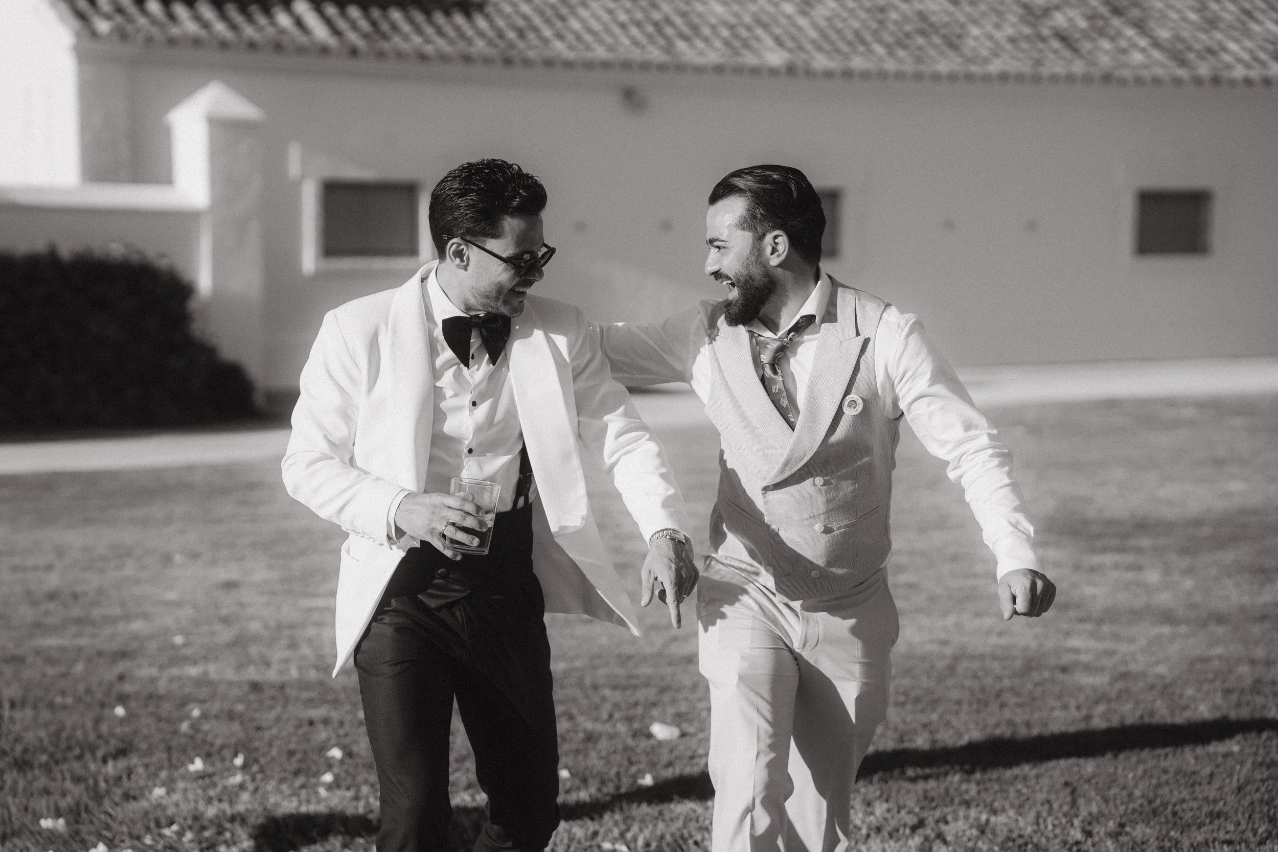 El novio y su hermano vestidos formalmente, uno con traje blanco y gafas y el otro con un traje gris y la corbata desajustada, riendo y caminando juntos en un entorno exterior