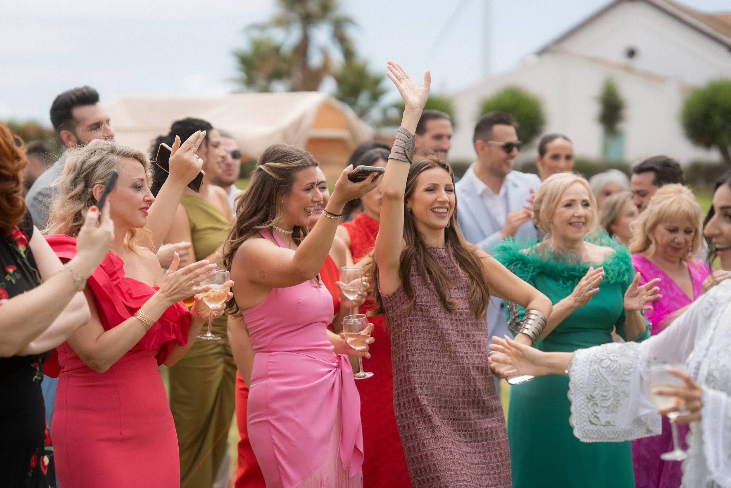 Grupo de personas en celebración, algunas con copas de vino o champagne, sonriendo y disfrutando del evento, con fondo de casas y árboles.