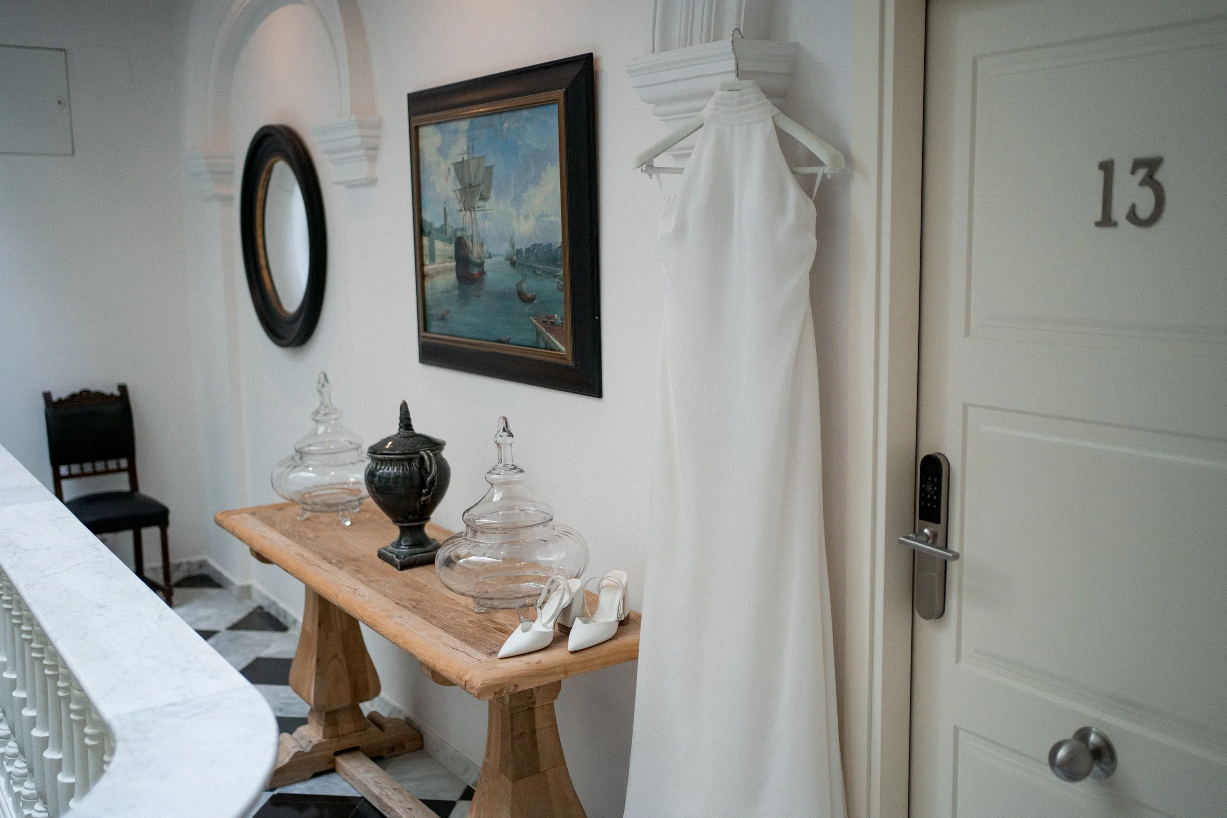 Vestido de novia blanco colgado en una percha en la pared, cerca de la puerta número 13 de un edificio, en un pasillo decorado con cuadros y objetos de cerámica sobre una mesa de madera. En el Hotel Armador en Cádiz.