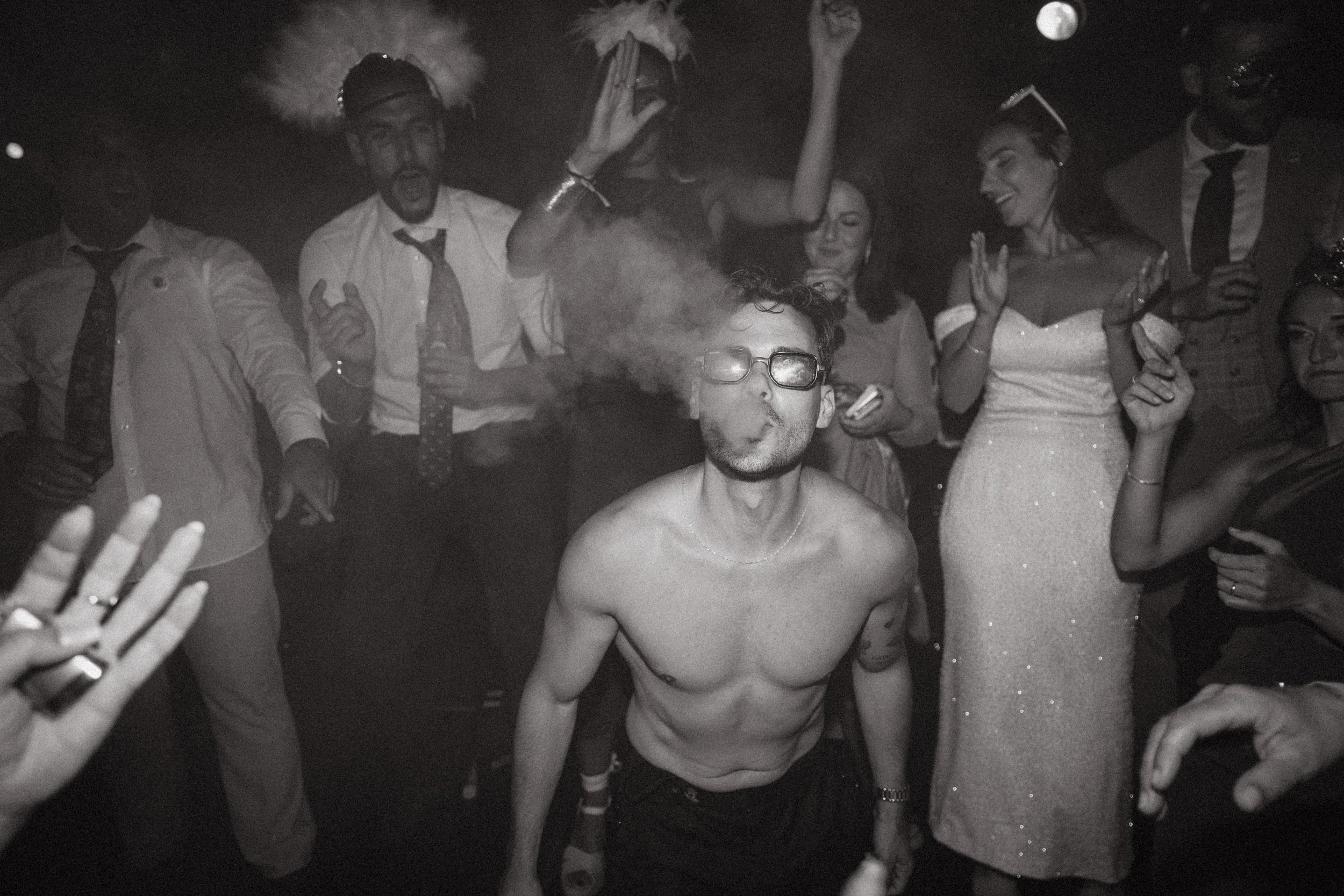Un grupo de personas en una fiesta, una persona en el centro está fumando y sin camisa, rodeada de amigos que celebran y disfrutan la noche.