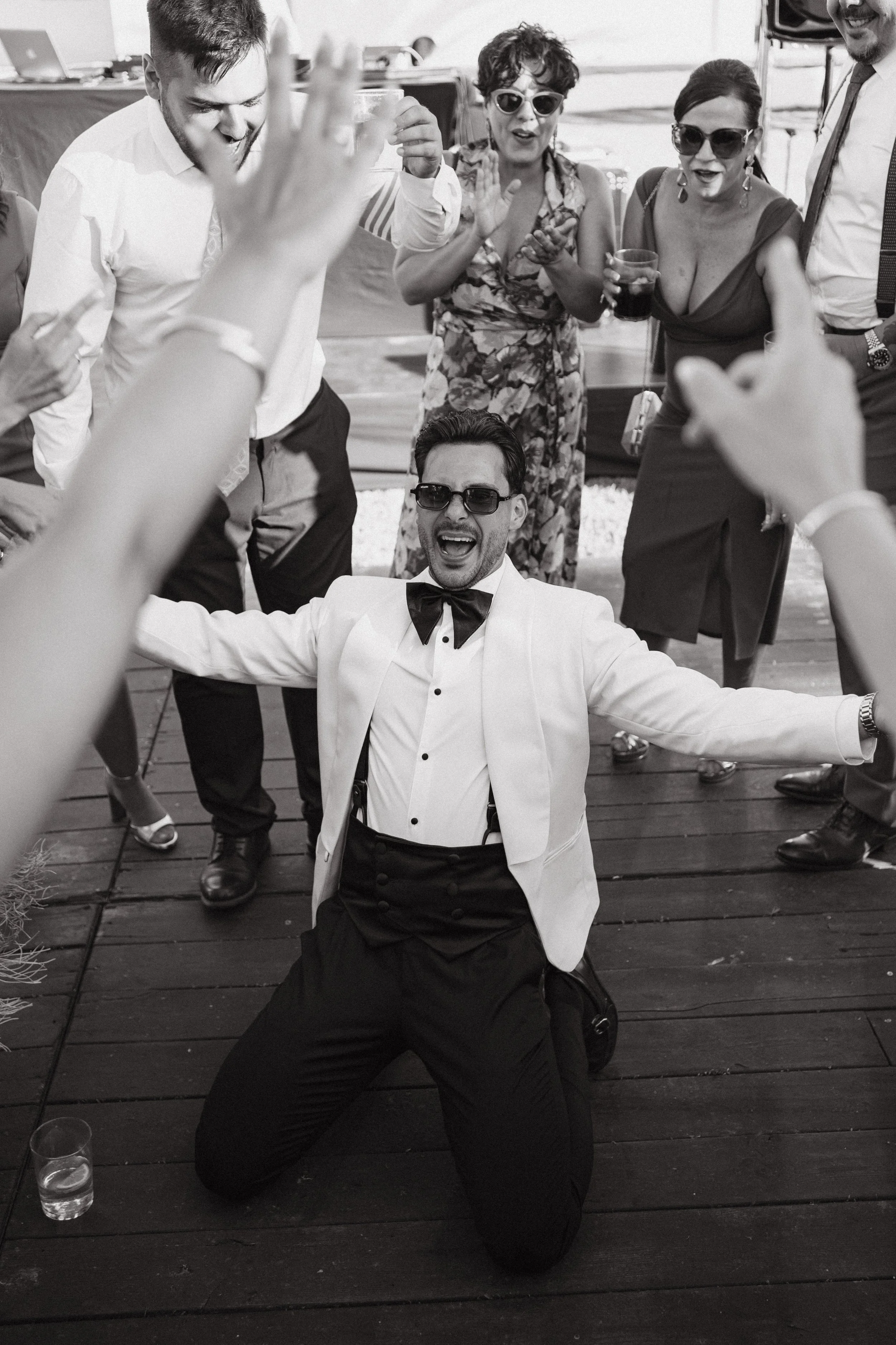 Grupo de personas bailando y disfrutando en una celebración, con un hombre en camisa blanca y vestido formal en el centro, rodeado de amigos, algunos con ropa elegante y gafas de sol, en un ambiente festivo al aire libre.
