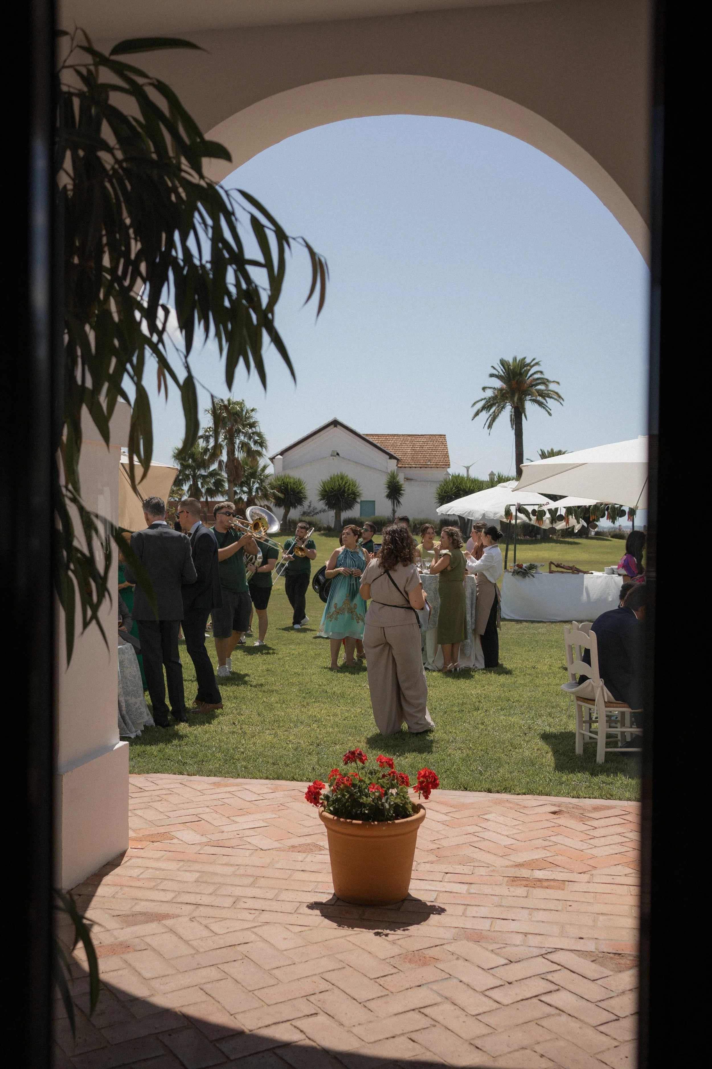 Vista de una boda al aire libre en Jerez de la Frontera, Cádiz. Con músicos tocando, personas conversando y decoraciones en un jardín con palmeras. En primer plano, una maceta con flores rojas sobre un piso de ladrillos, visto desde un arco.