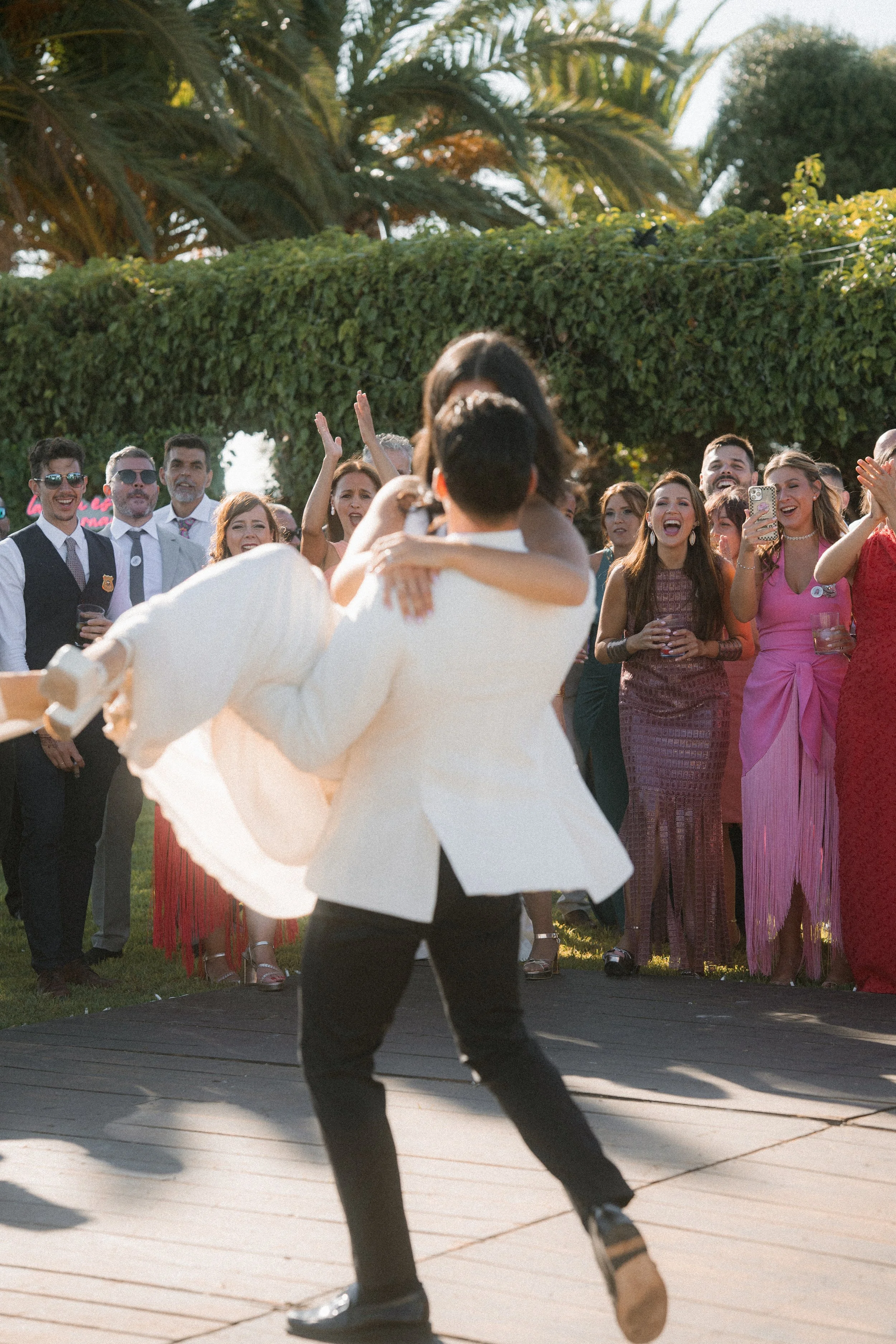 Pareja bailando en una boda al aire libre con invitados sonrientes y aplaudiendo en el fondo.