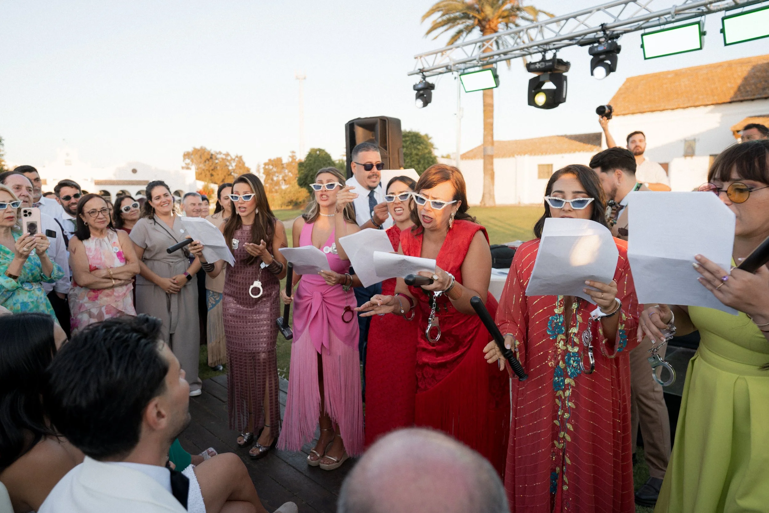 Grupo de personas cantandole una tanda de cuplés a los novios, algunos con gafas de sol y sosteniendo cupletinas, mientras otros observan, en un evento al aire libre con iluminación de escenario.
