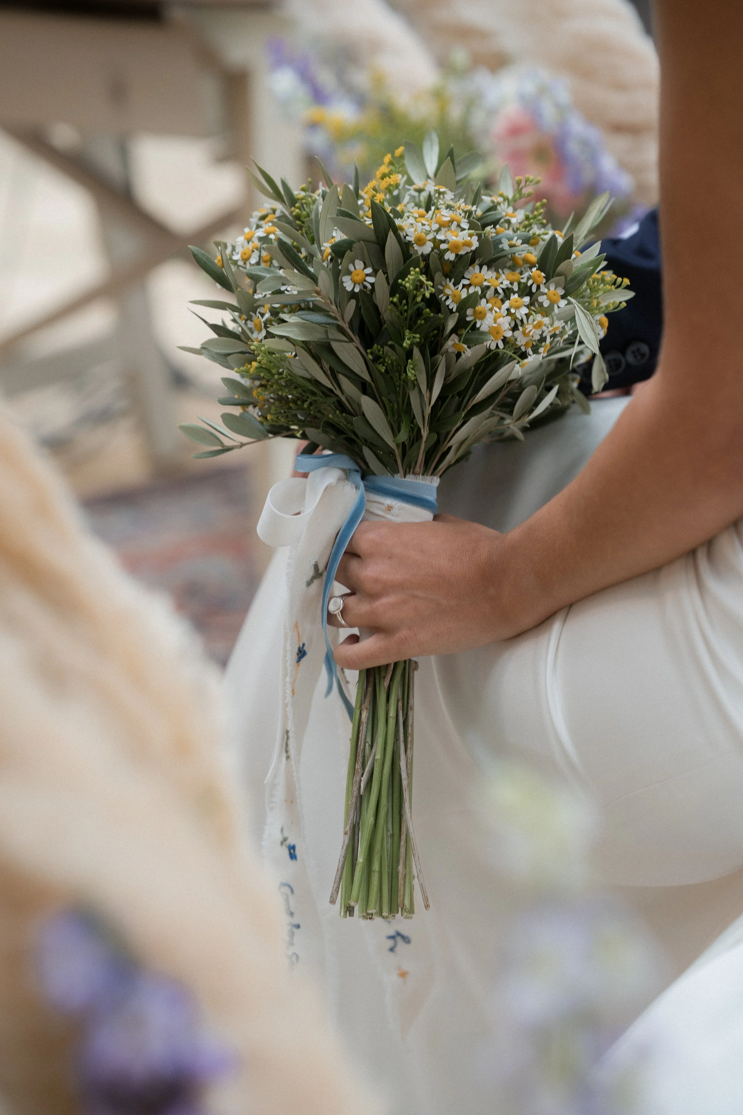 Una persona sostiene un ramo de flores con margaritas y hojas verdes, adornado con cinta blanca y azul.