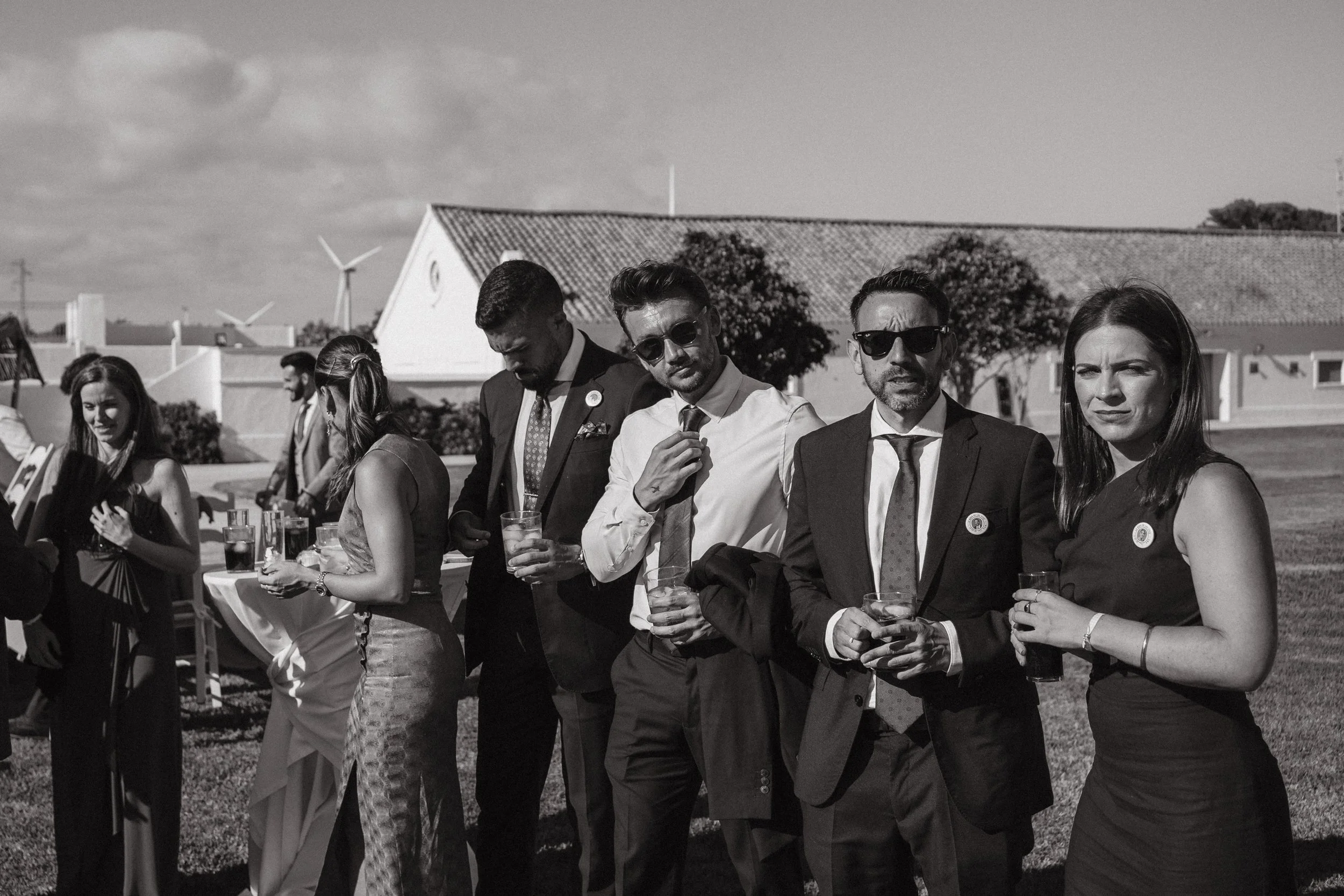 Grupo de personas en evento formal al aire libre, sosteniendo bebidas, vestimenta elegante, algunas con gafas de sol, en un entorno con césped y edificios al fondo, en blanco y negro.