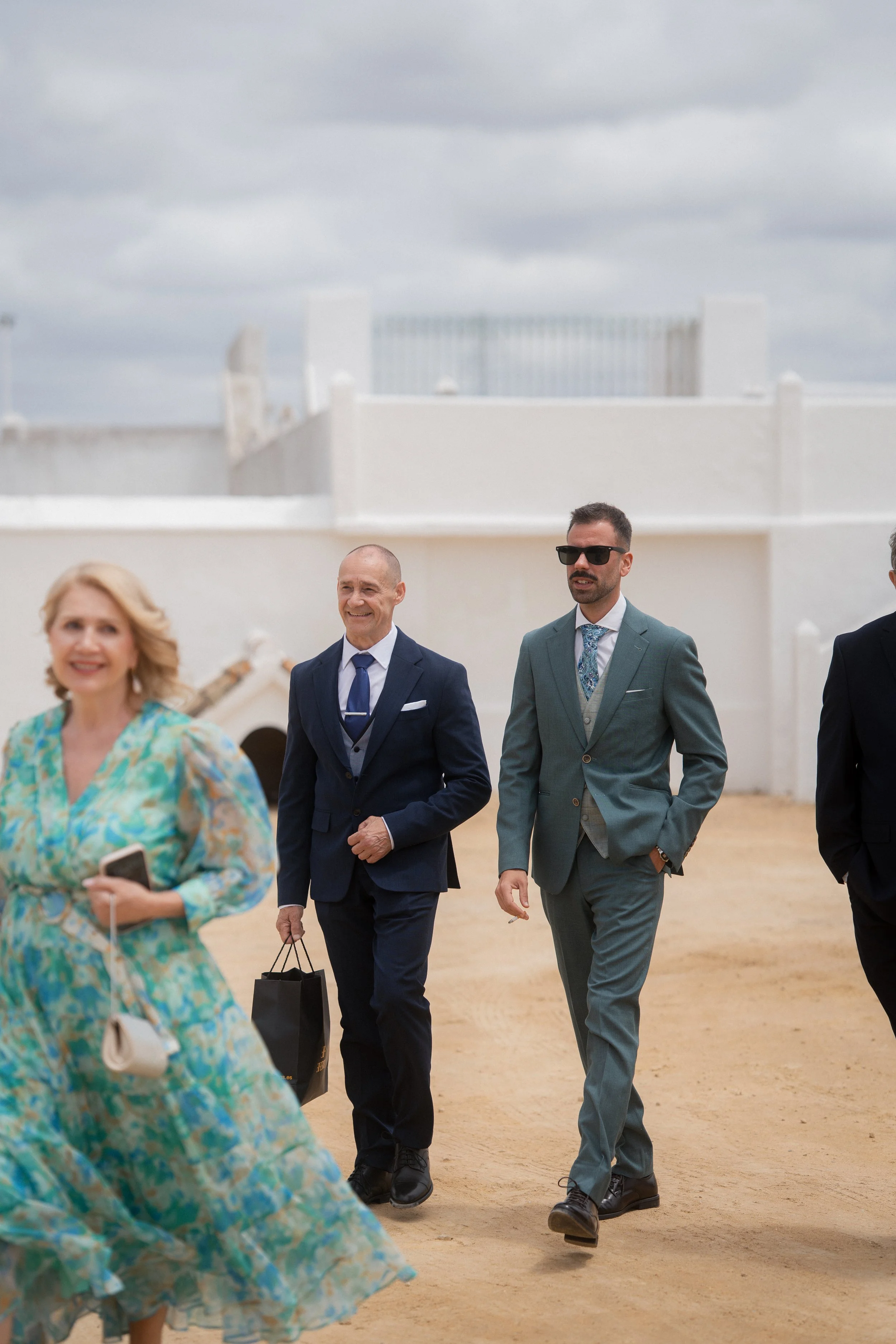 Personas caminando en una especie de evento al aire libre en un día nublado, con construcciones blancas de fondo.