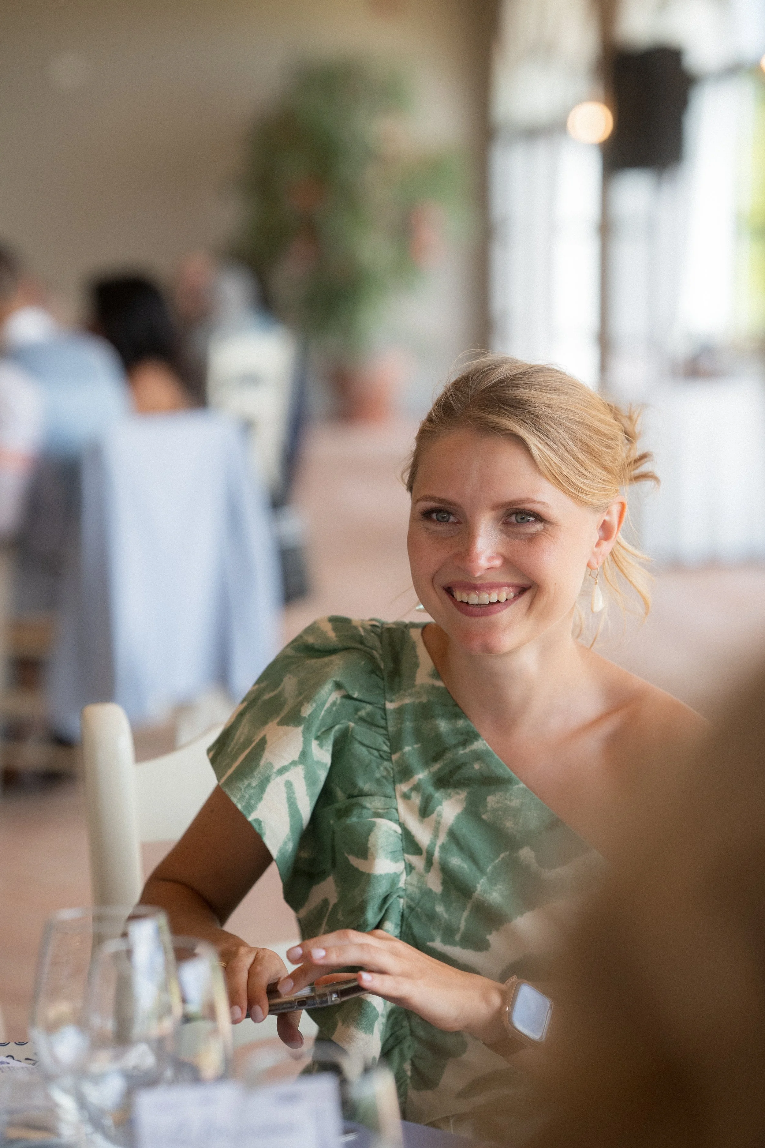 Mujer sonriendo en una reunión social en un restaurante o salón de eventos, con fondo difuso de otras personas y decoraciones.