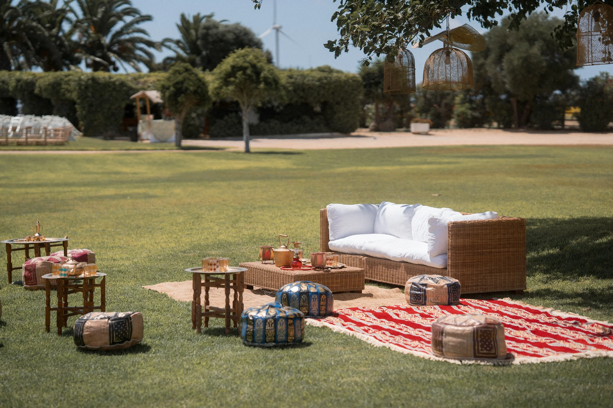 Una sala de picnic en un césped con un sofá de canasta, mesas pequeñas, cojines, una alfombra roja y teteras. Rodeado de árboles y arbustos en un día soleado.