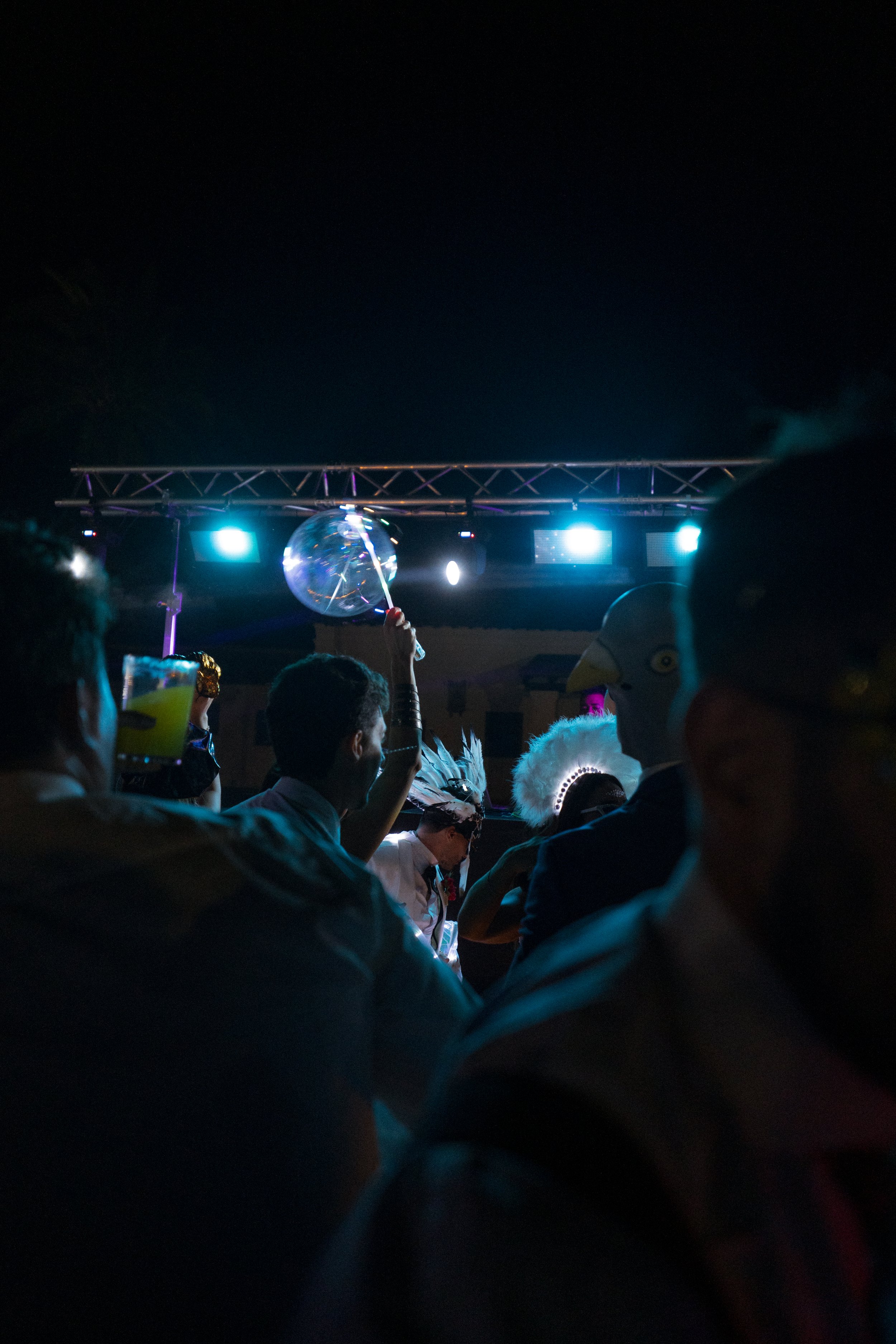 Personas en una boda en exterior por la noche con iluminación azul y un escenario, algunos con disfraces con plumas y máscara, y una persona con un gorro con forma de pájaro que sostiene un globo transparente.