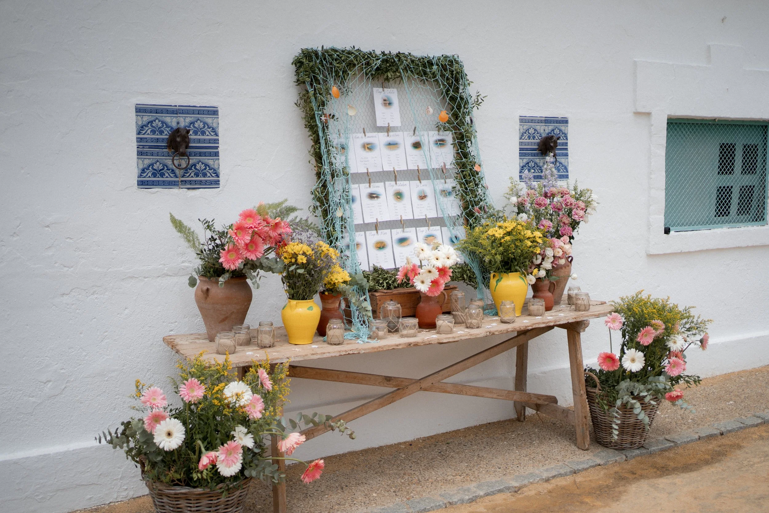 Mesa de madera con arreglos de flores en jarrones de cerámica y cestos, decorada con una rama pintada de azul y papel colgado, en un fondo de pared blanca con ventanas y azulejos decorativos.