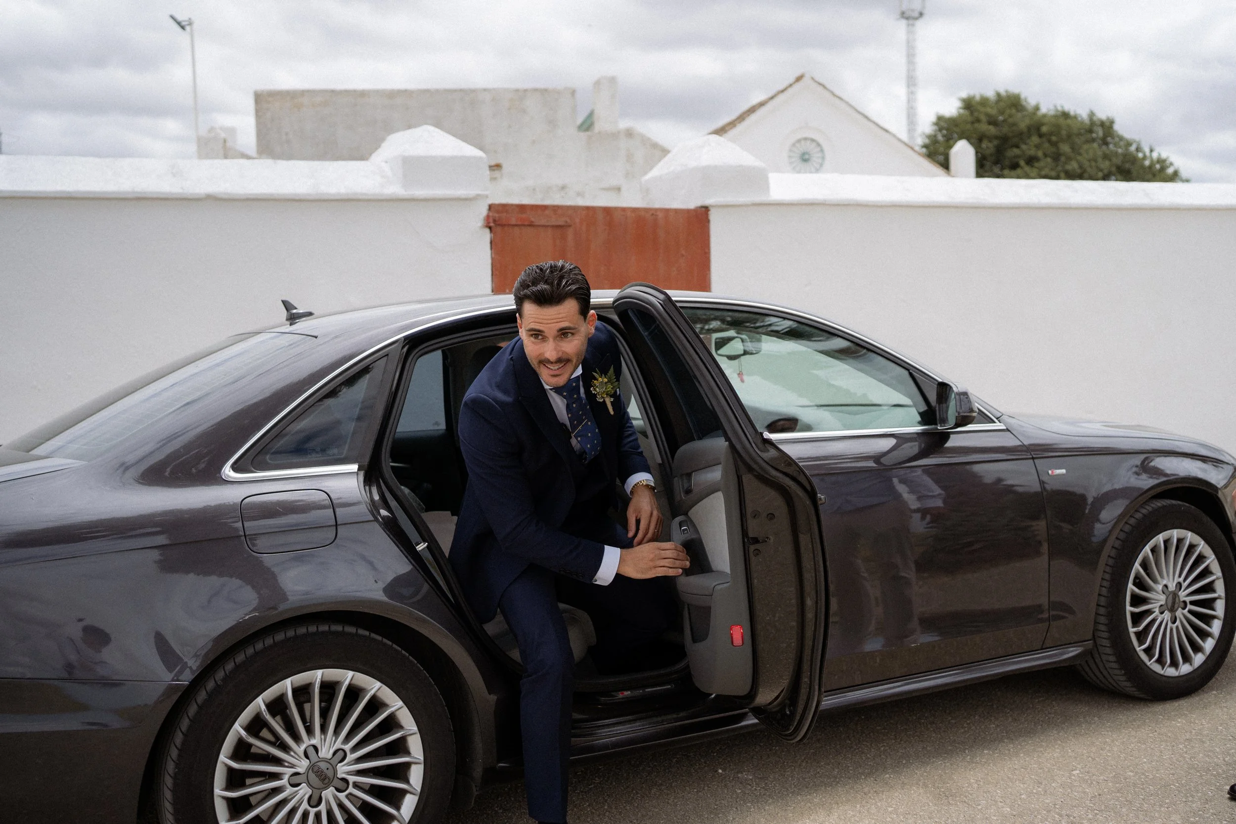 Hombre vestido con traje entrando en un coche negro desde la puerta trasera, en un entorno exterior con paredes blancas y un edificio con techo de tejas.