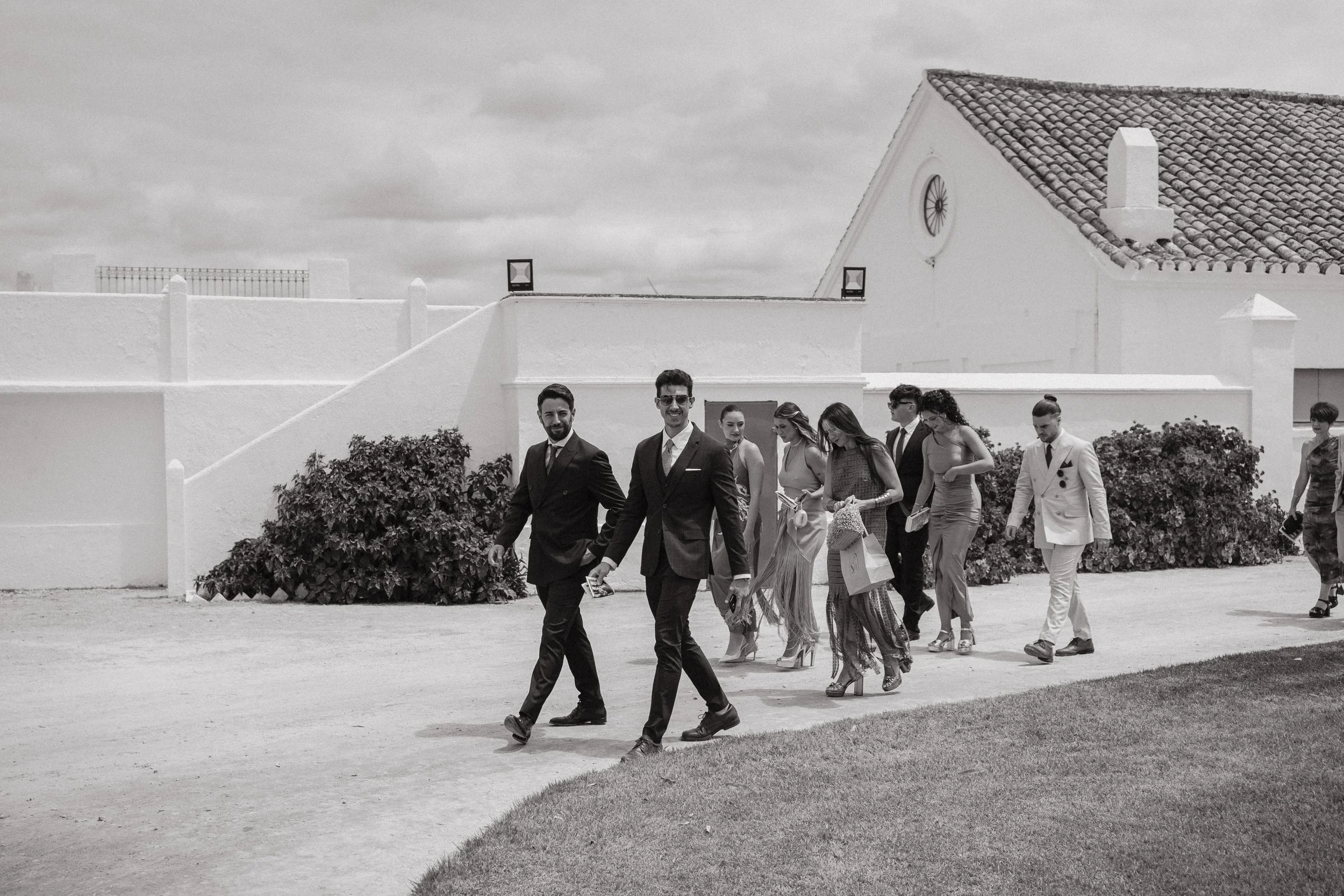 Grupo de personas vestidas formalmente, caminando en un exterior con un edificio de techado de tejas en el fondo.