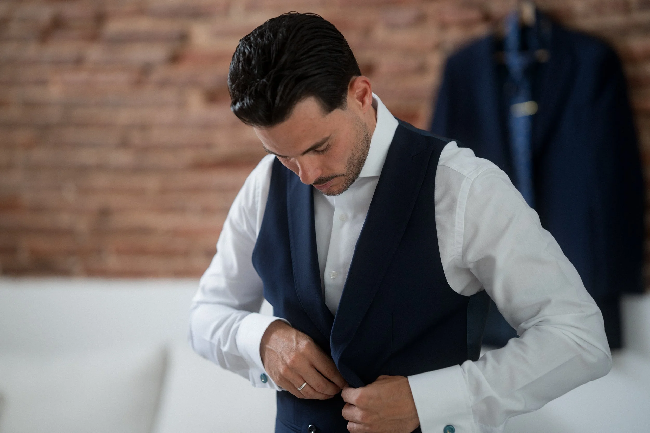 Hombre con peinado oscuro y barba ajustando su chaleco y camisa blanca en un espacio con pared de ladrillos y ropa colgada al fondo.