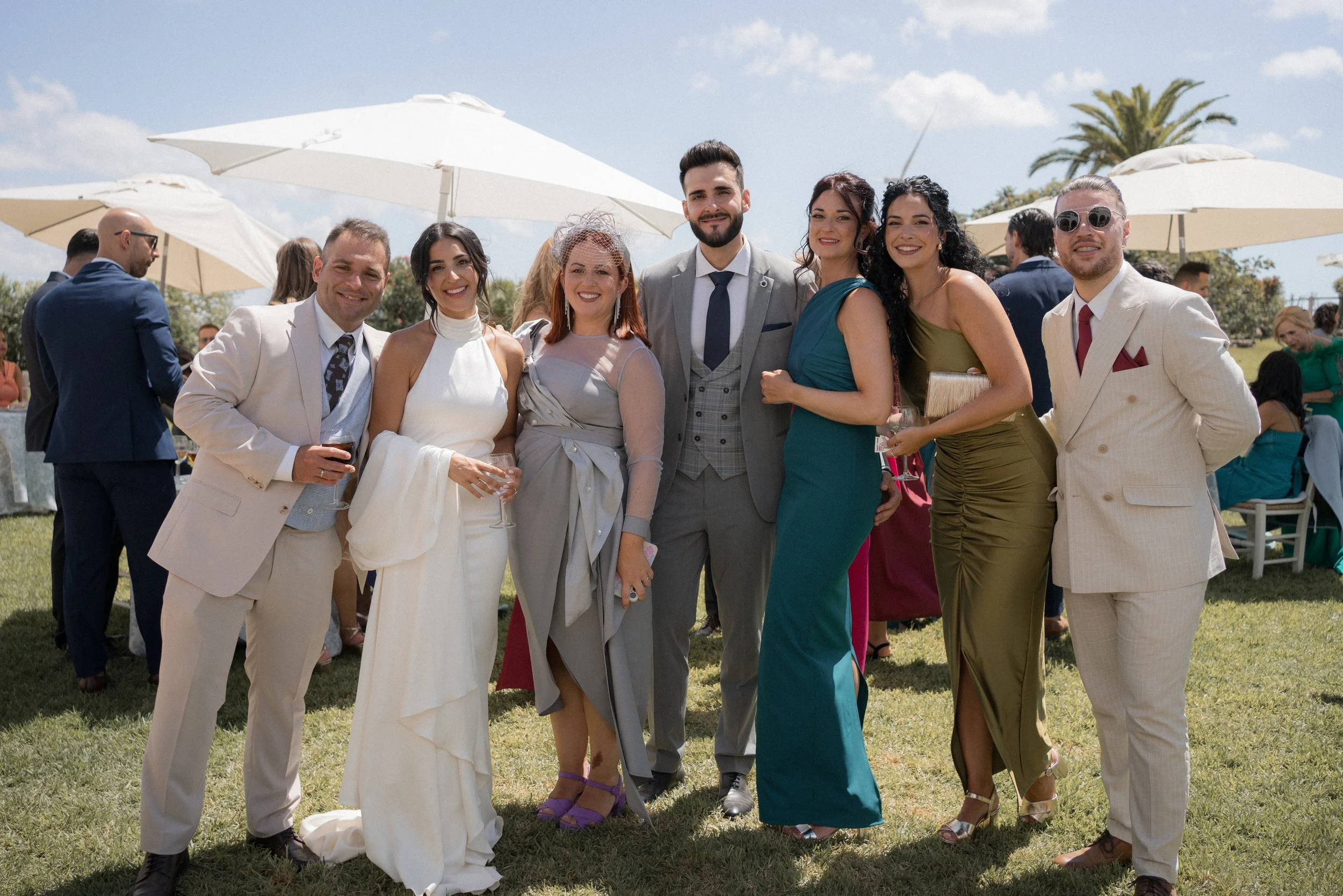 Grupo de personas en una celebración al aire libre, vestidas con ropa elegante, en un día soleado con sombrillas y palmeras de fondo.