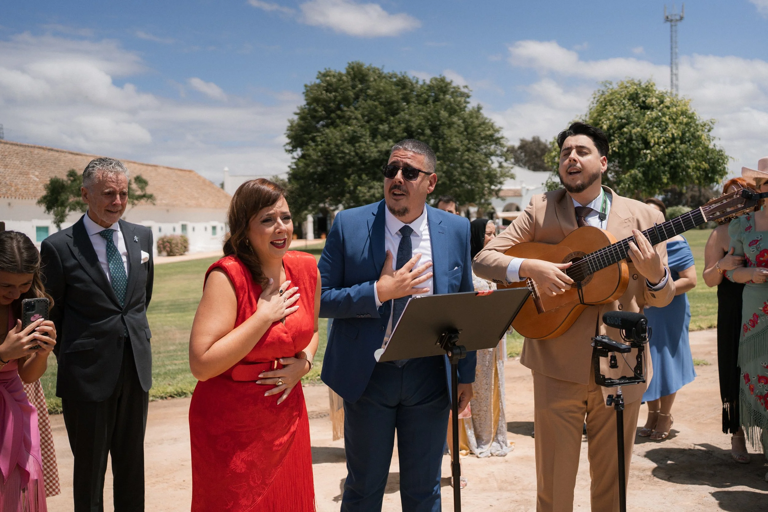 Grupo de personas en una ceremonia al aire libre, cantando y tocando guitarra, con fondo de cielo y árboles.