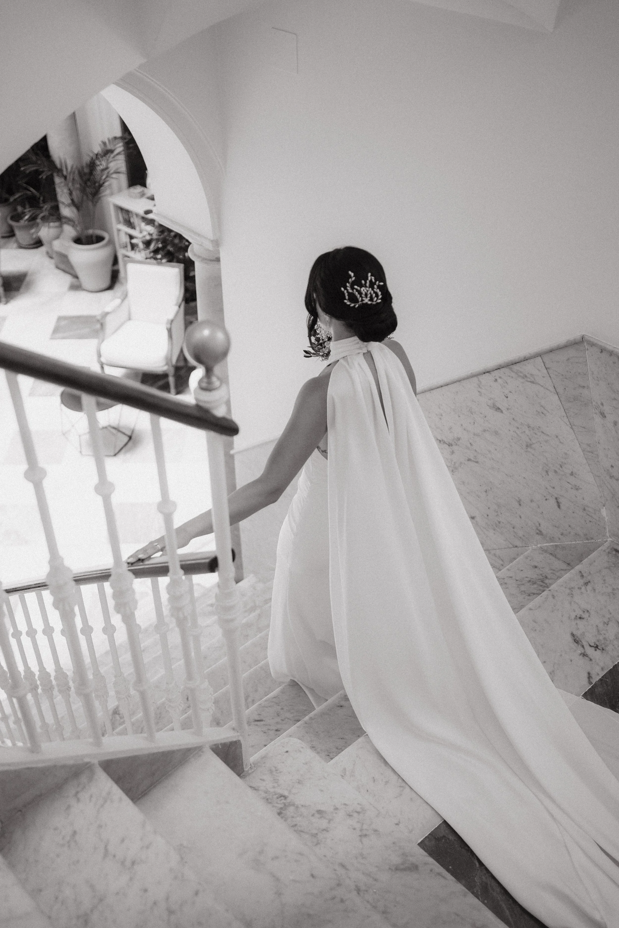 Mujer vestida con un vestido blanco largo, con un velo y accesorios en el cabello, bajando una escalera de mármol en un interior decorado con plantas y muebles.
