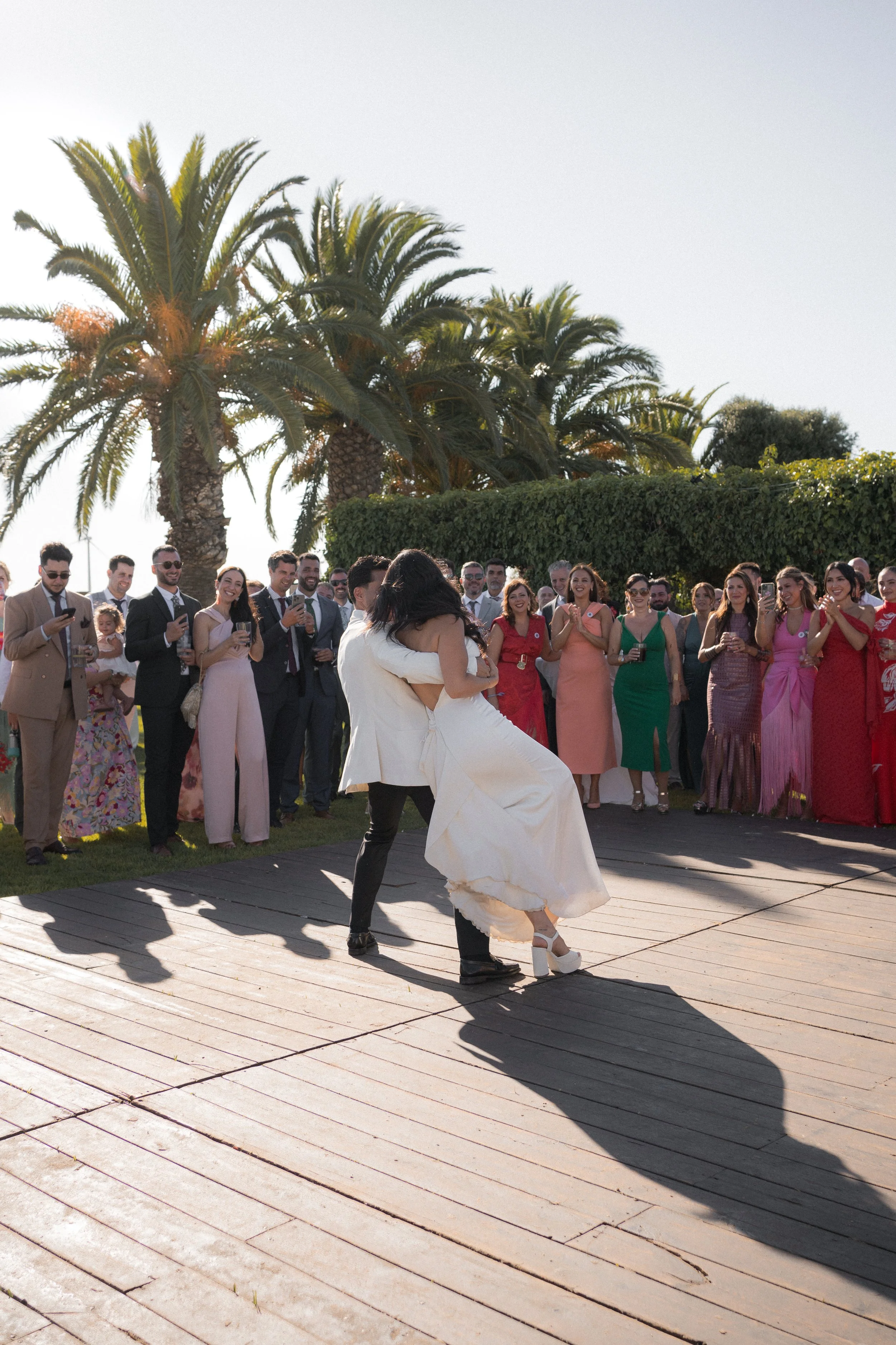 Una pareja bailando en una ceremonia de boda al aire libre, rodeada de invitados sonriendo y tomando fotos, con palmeras y vegetación en el fondo.