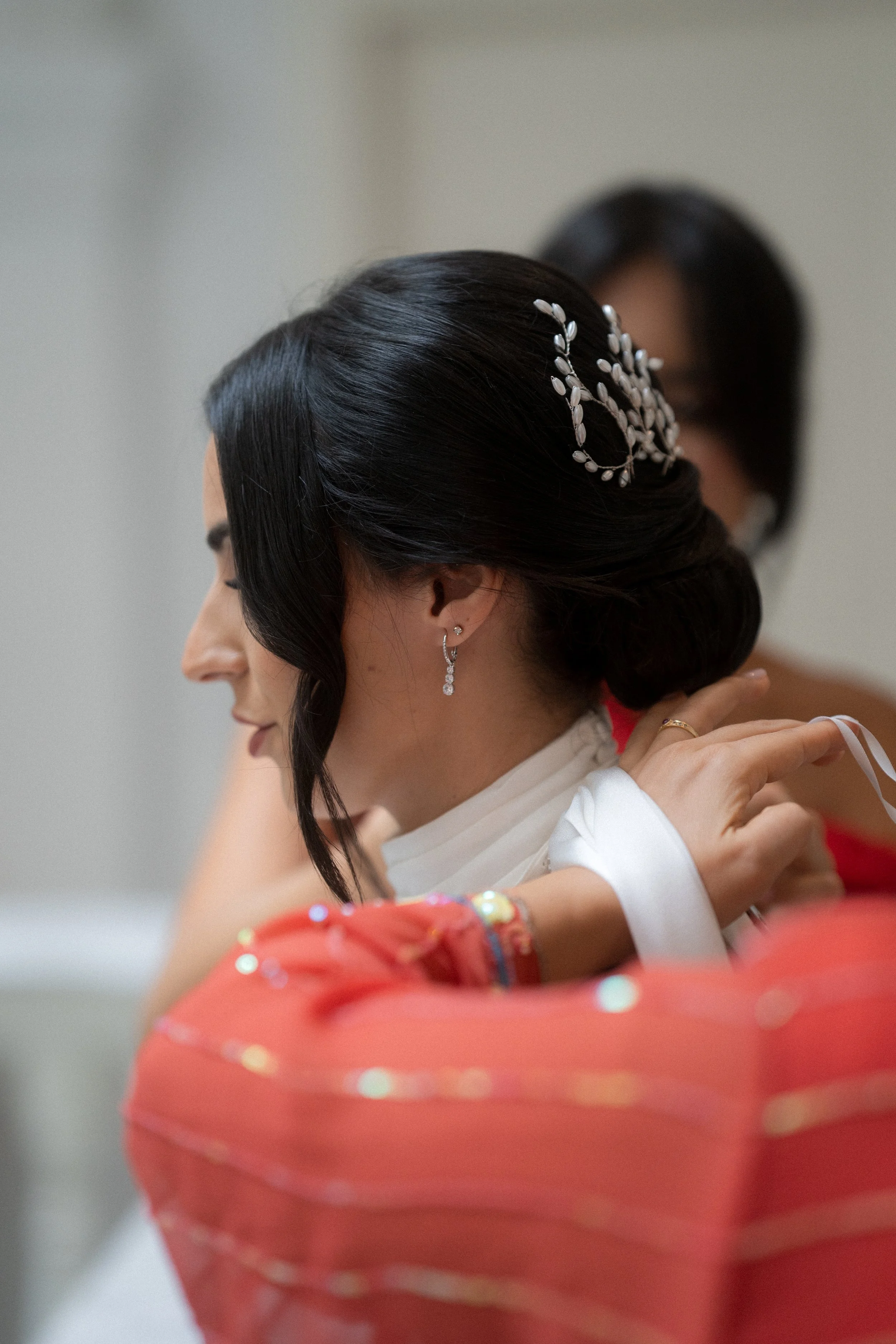 Mujer con cabello oscuro y peinada en un estilo elegante, usa aretes y adornos en el cabello, vestida con ropa blanca y rodeada de otras personas en un entorno formal.