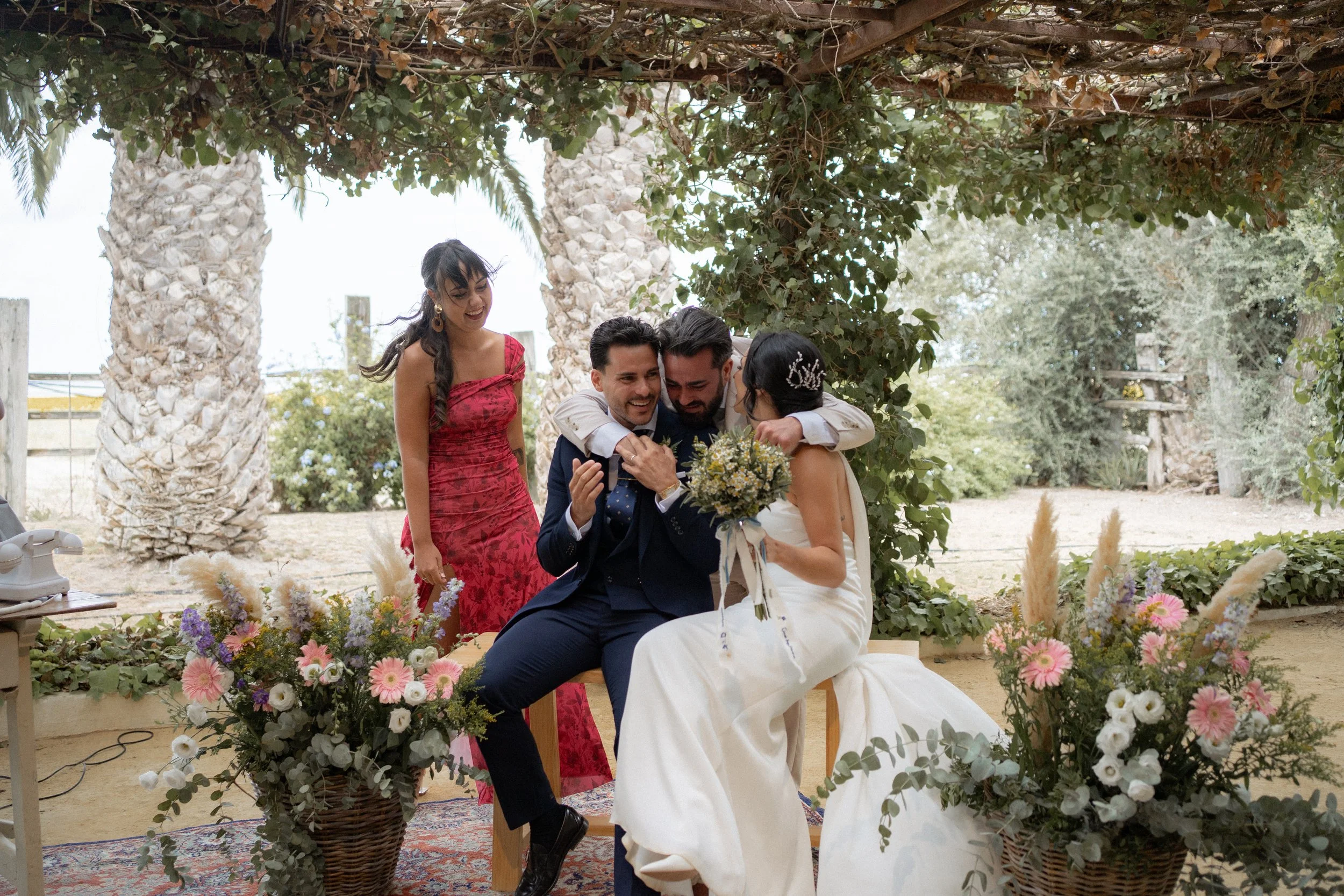 Una ceremonia de boda al aire libre con cuatro personas, donde una pareja está sentada en una banca con la novia sosteniendo un ramo de flores y los amigos celebrando emocionalmente. Hay decoraciones florales y vegetación alrededor.